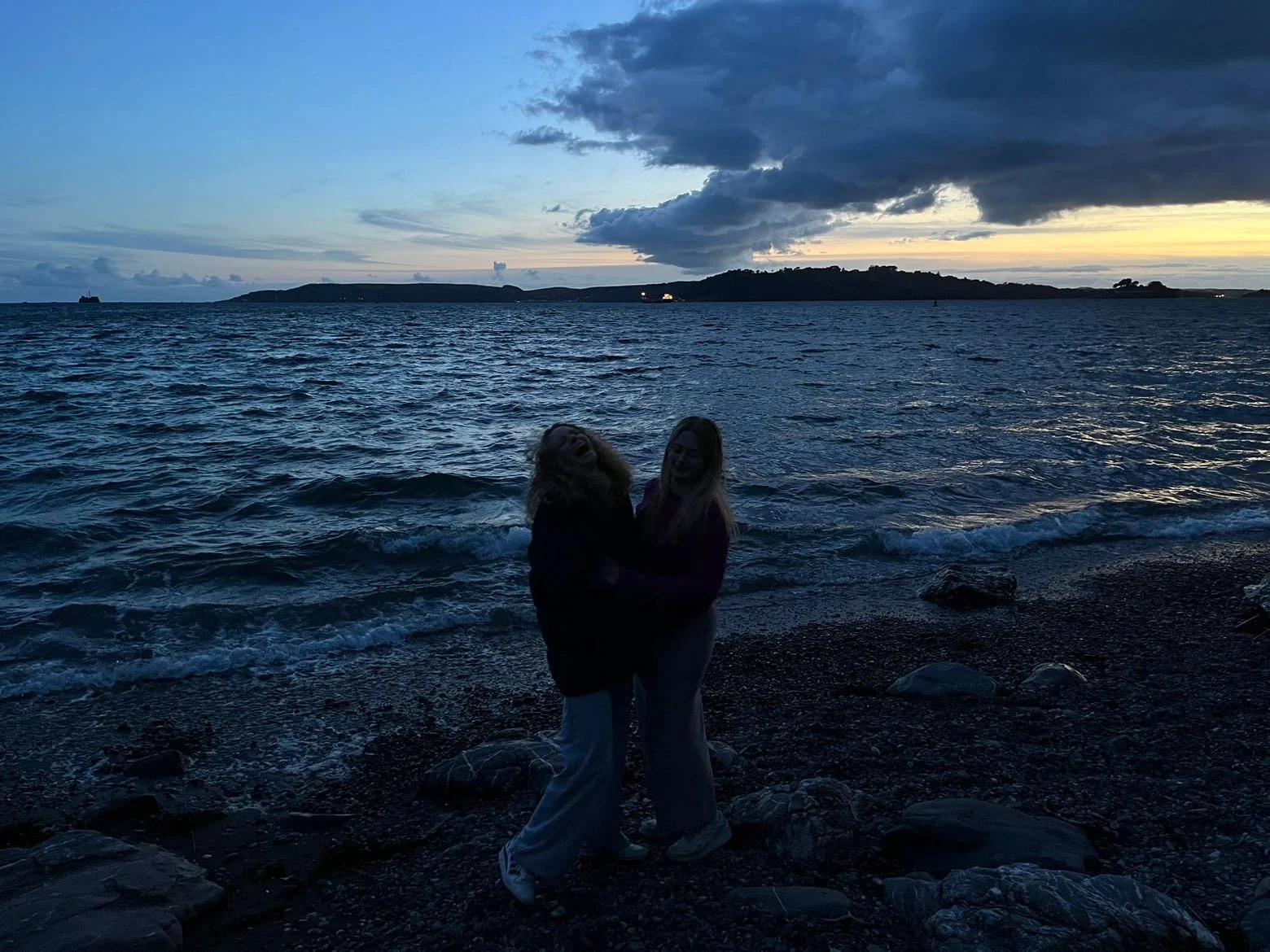 Two people standing on a rocky beach at sunset, silhouetted against the ocean and sky with dark clouds and distant land.