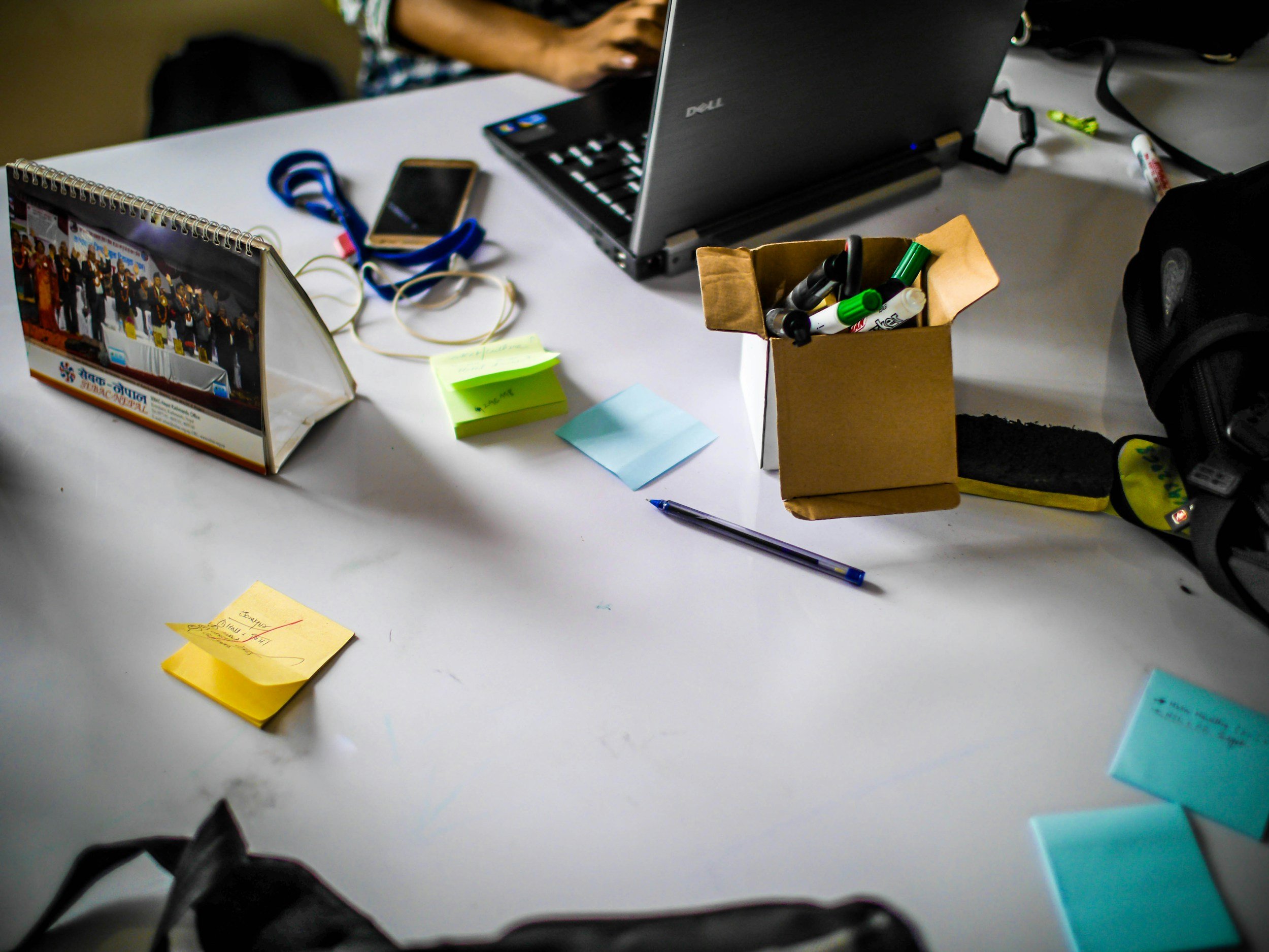 A cluttered desk with a laptop, sticky notes, pens, markers, a smartphone, a calendar, and a backpack.
