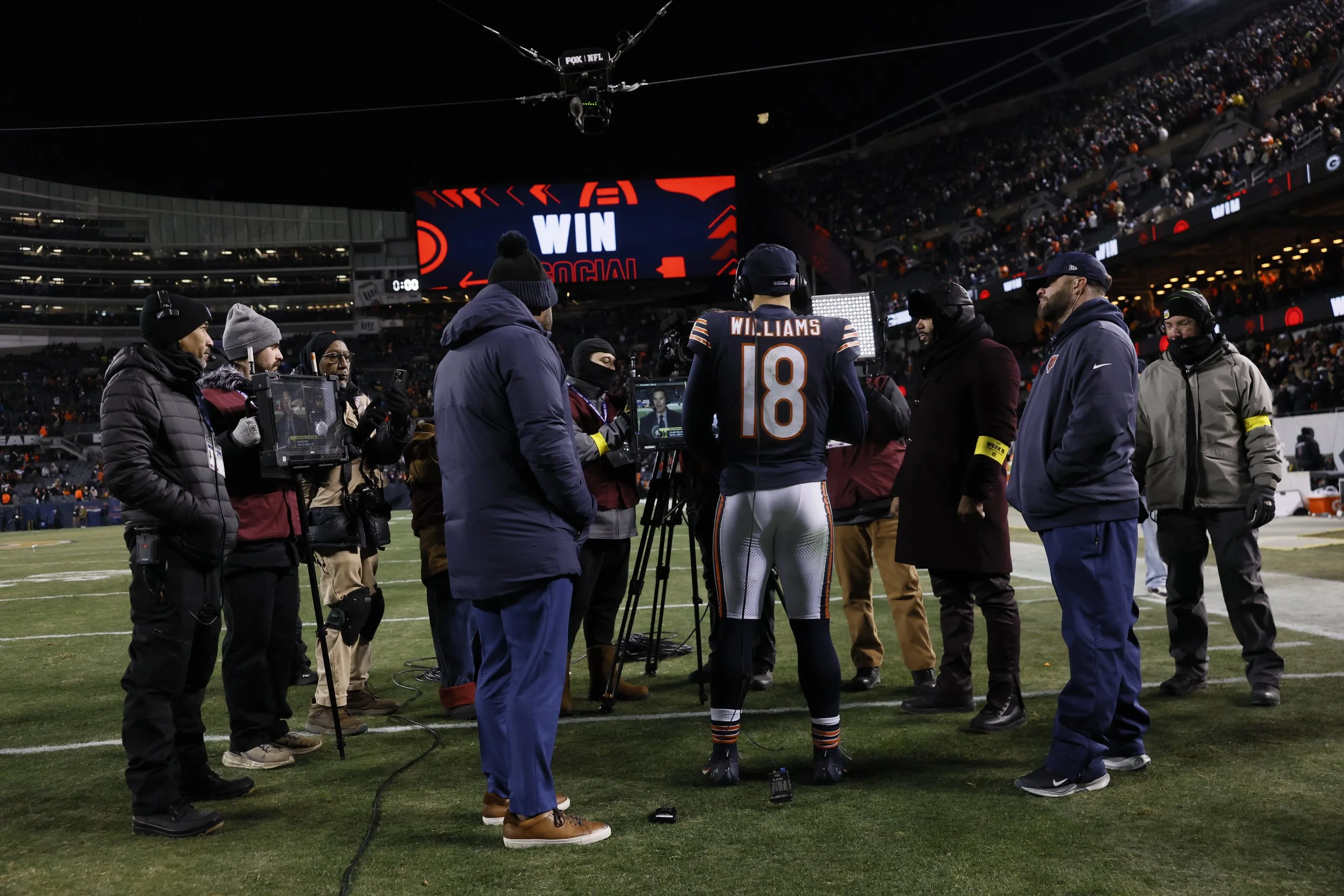 A football player in uniform with number 18 and the name Williams on the back is conducting interviews or media coverage after a game, surrounded by media personnel and camera operators on a football field at night with a large screen displaying "WIN" and "SOCIAL" in the background.