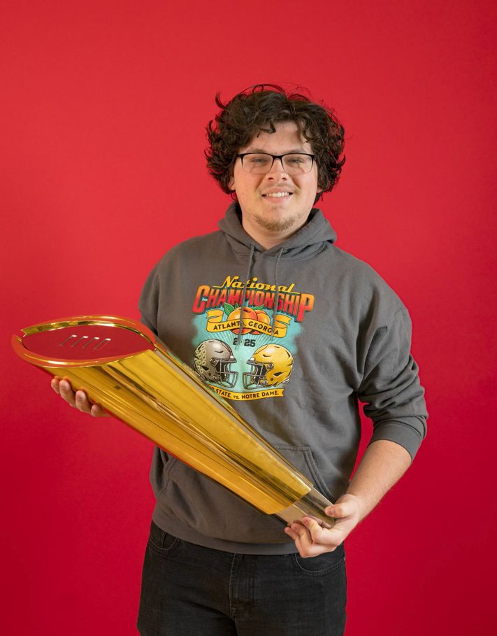 A young man with curly hair and glasses holding a large golden trophy in front of a red background.