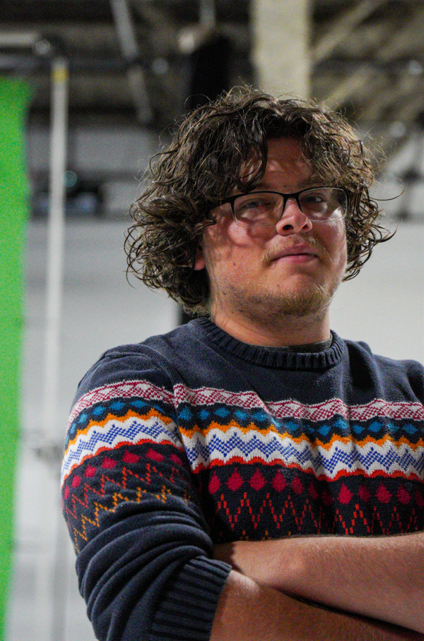 A young man with curly hair and glasses, wearing a colorful patterned sweater, standing with arms crossed indoors.