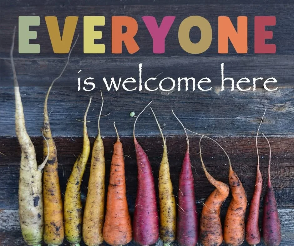 Colorful carrots arranged in a row with a sign that reads 'Everyone is welcome here' above them.