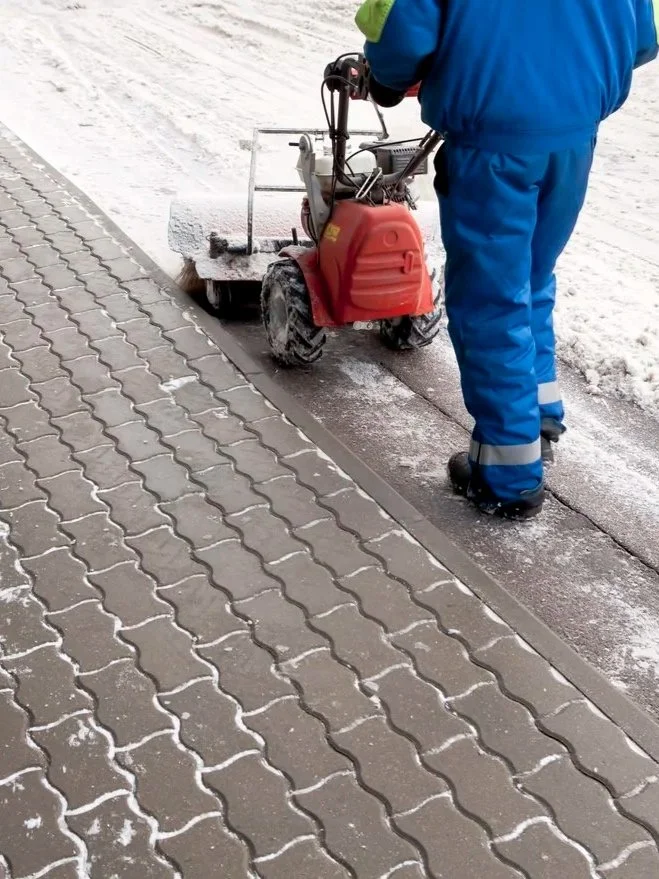 Worker in blue winter coat and pants using a machine to remove snow or ice from a sidewalk.