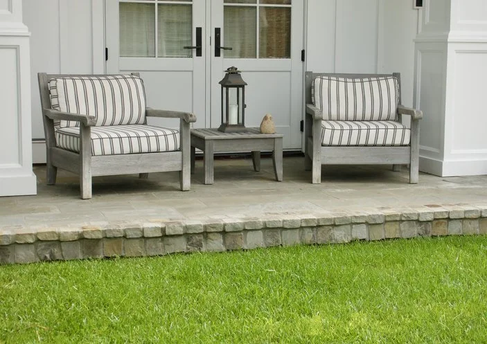 Two outdoor wooden chairs with striped cushions, a small wooden table with a lantern and decorative item, on a porch with glass-paneled doors and a lawn in the foreground.