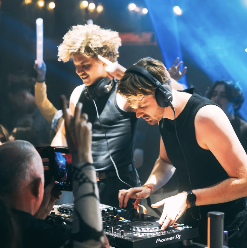 Two male DJs performing at a nightclub, one with curly blond hair and the other with straight brown hair, both wearing headphones, with a crowd taking pictures in front of them and colorful stage lighting.