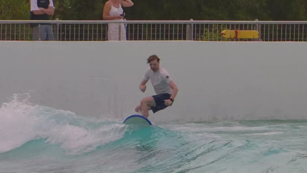 A young man surfing on a wave at a water park or pool with a white wall and onlookers in the background.