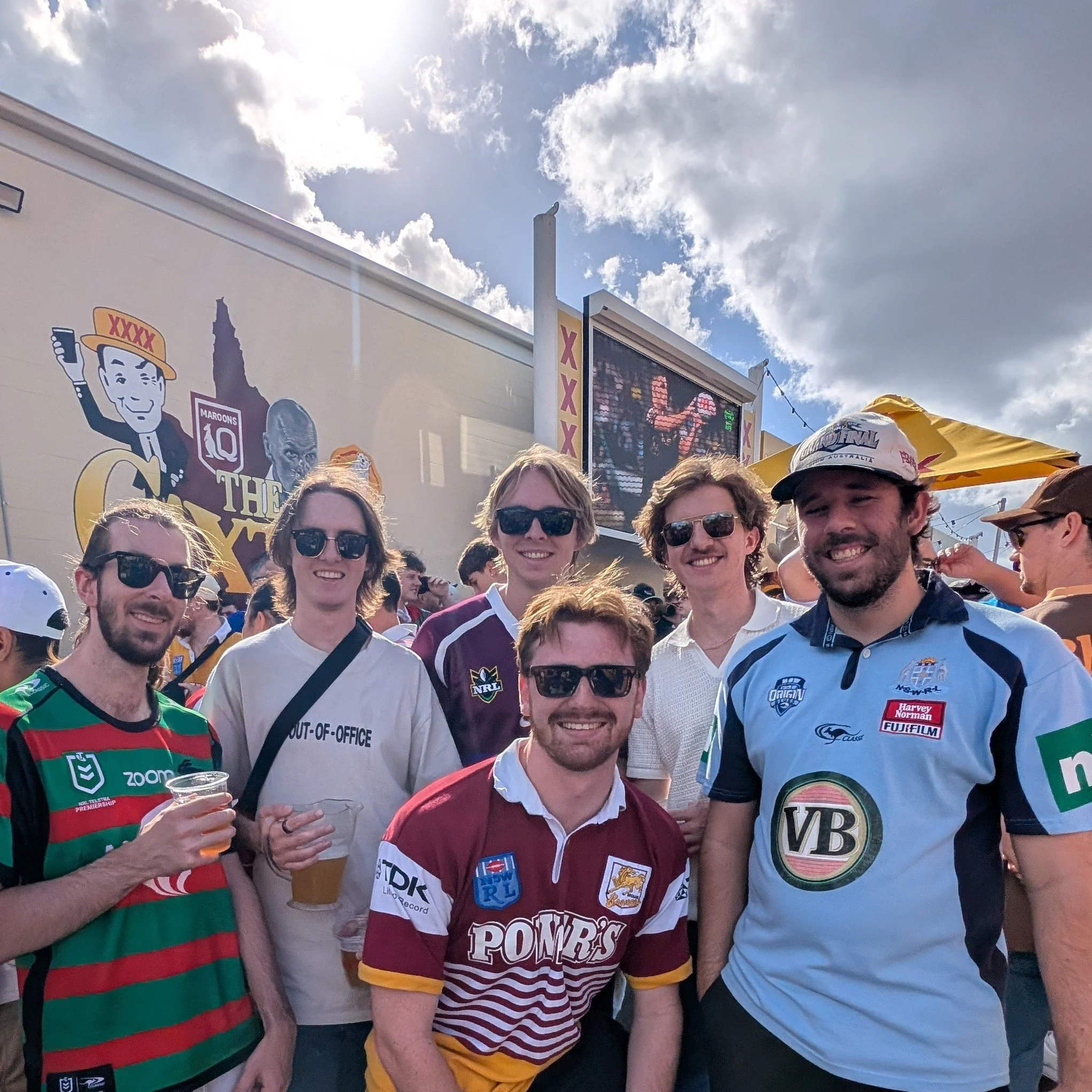 Group of friends at an outdoor event, smiling at the camera, with a large screen and advertising banners in the background, including a cartoon of a man with a drink and a silhouette of a person, under partly cloudy skies.