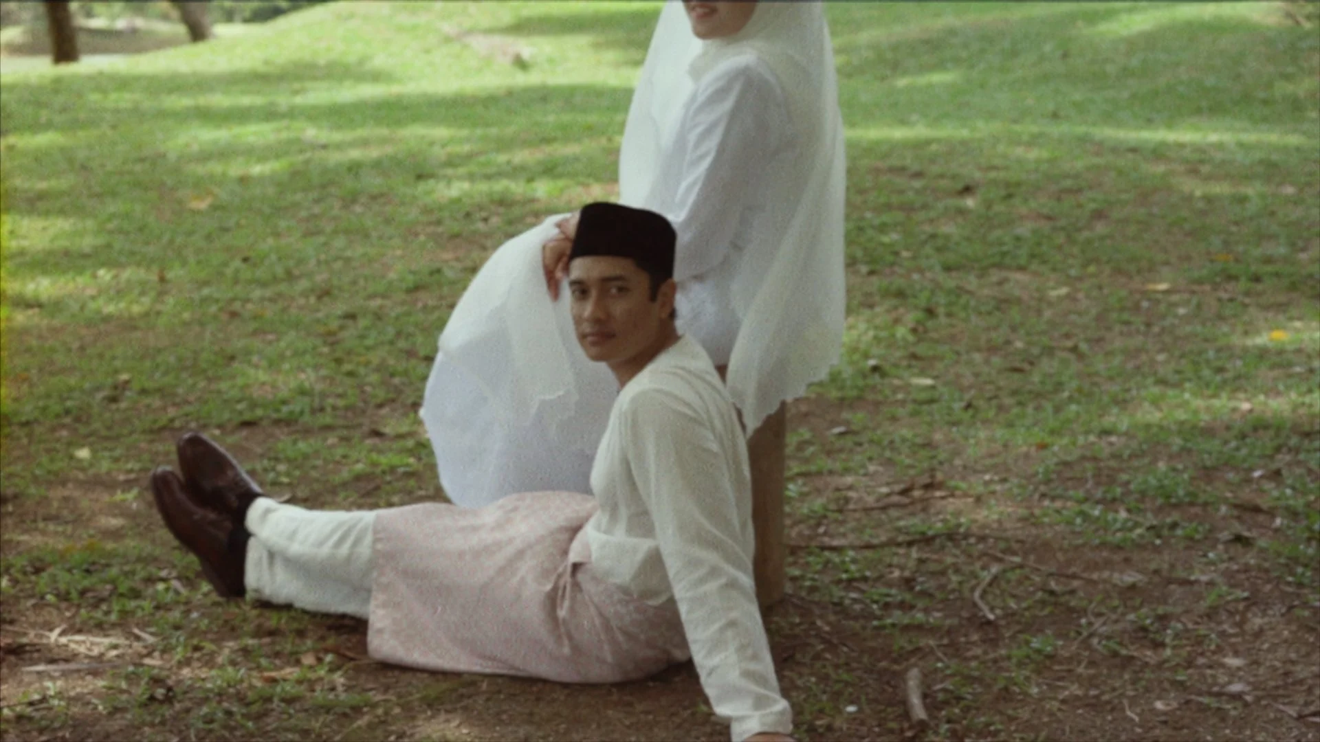 Two men dressed in white traditional clothing, one sitting on the ground and the other leaning on him with a hand on his head, outdoors on grass under trees.
