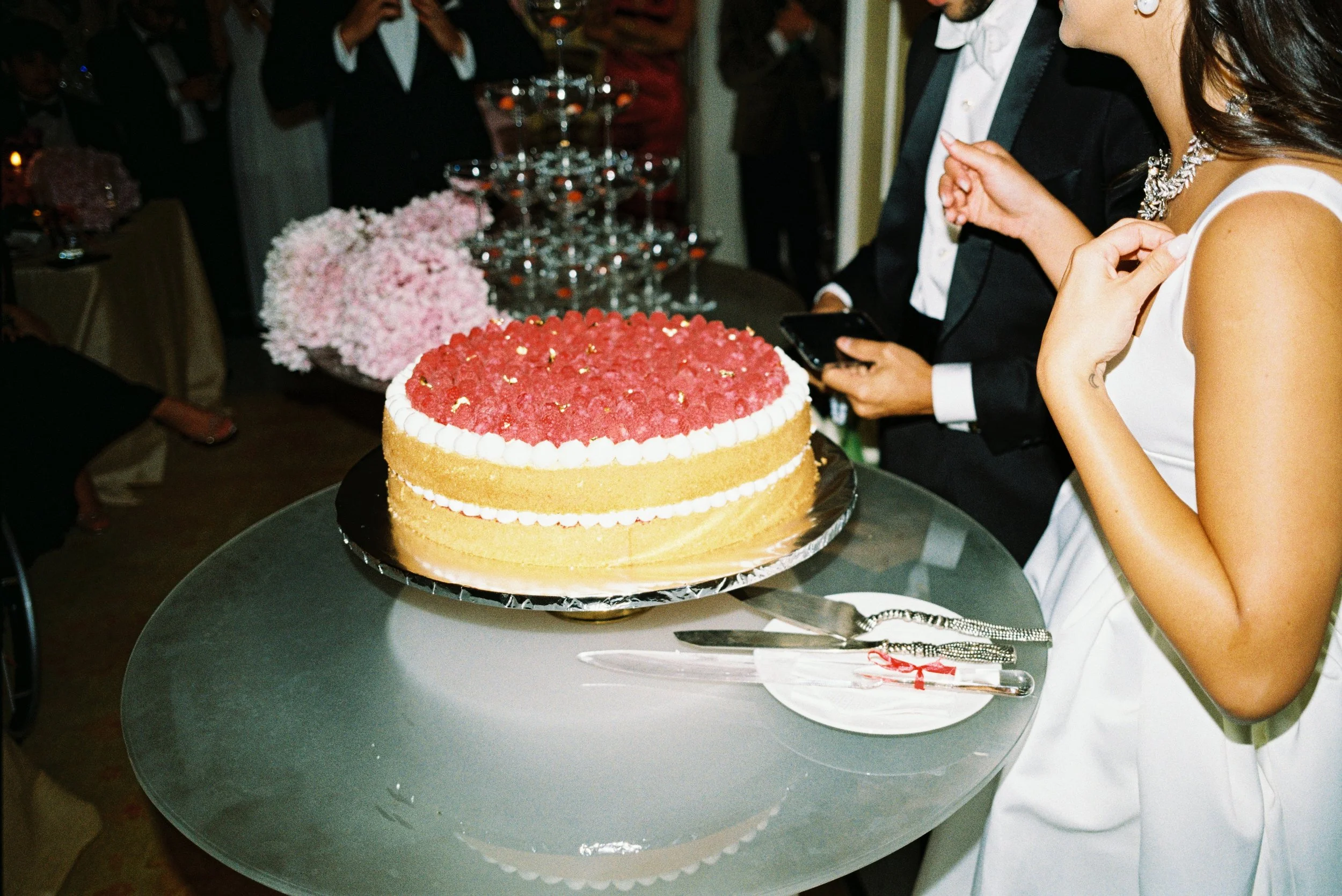 A layered birthday cake with pink icing on top and white piped borders, placed on a silver cake board on a round table, with people in formal attire celebrating in the background.