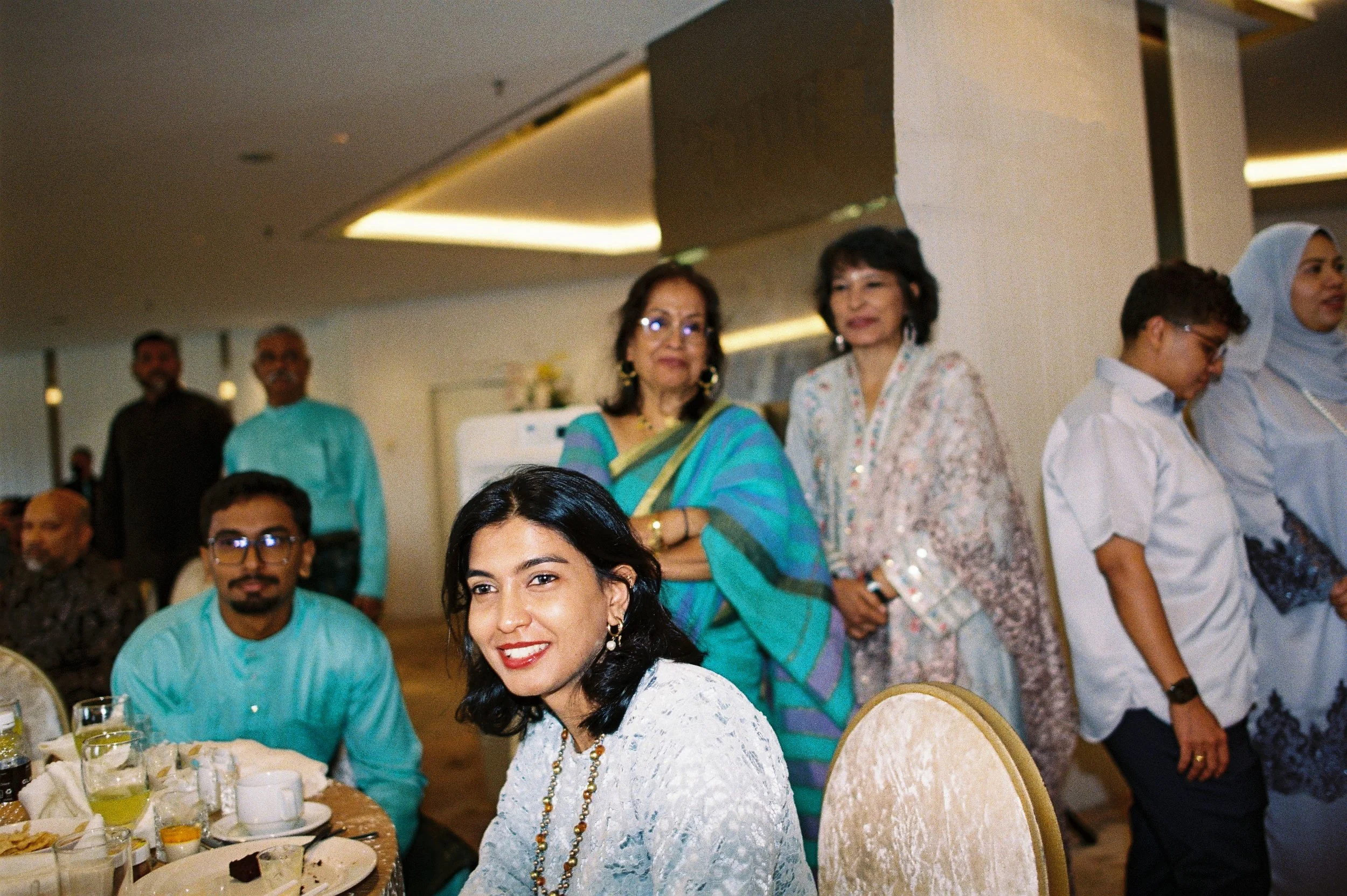A group of people gathered at a social event, with women standing and sitting around a table set with glasses and plates, dressed in traditional and formal attire.