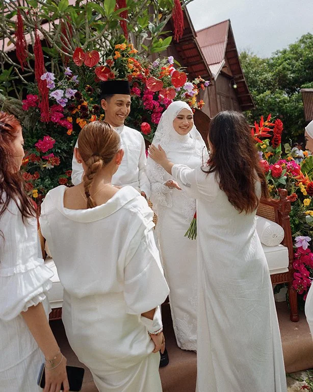A bride and groom dressed in white traditional wedding attire smiling and greeting friends at a wedding ceremony outdoors, surrounded by colorful flowers and greenery.
