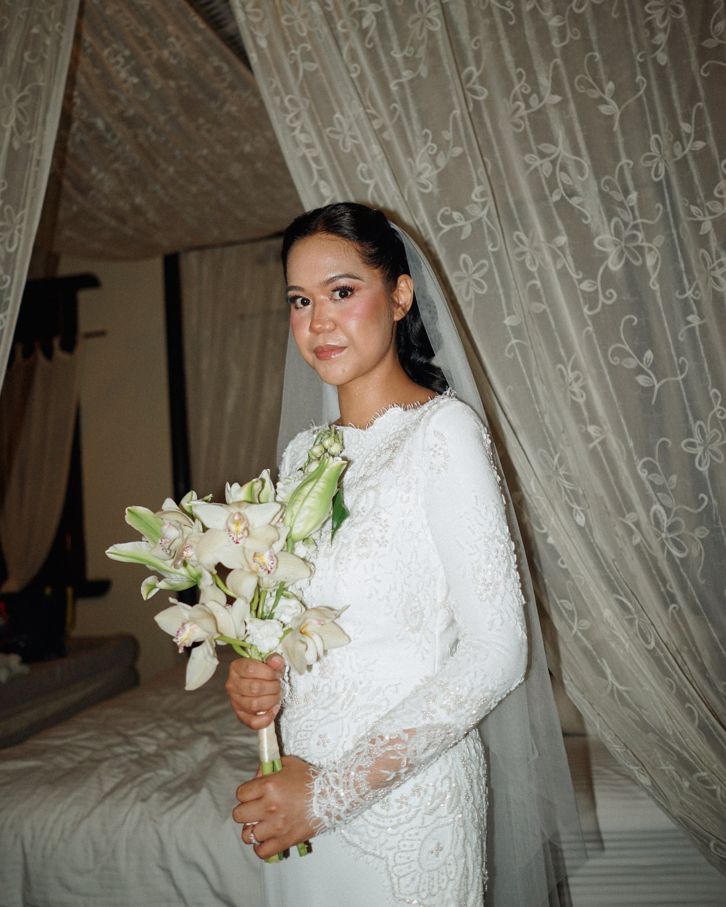A bride in a white wedding dress holding a bouquet of white flowers, standing in a room with lace and floral curtains.