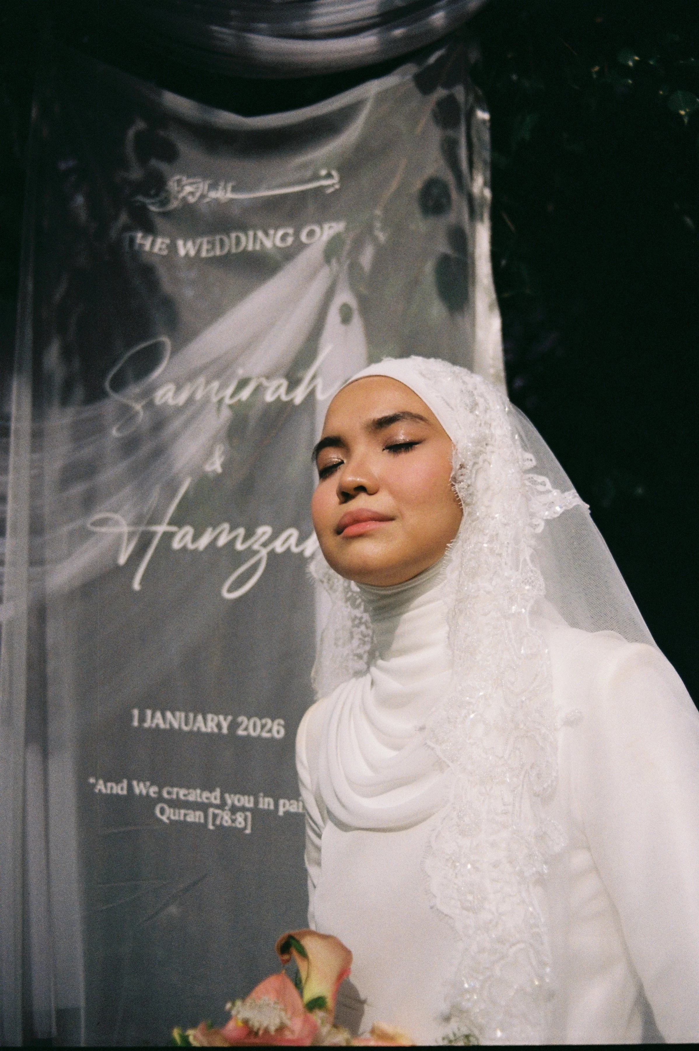 A woman in a white wedding dress with lace details and a hijab, with her eyes closed, standing in front of a black backdrop that has white text about a wedding date in January 2026.