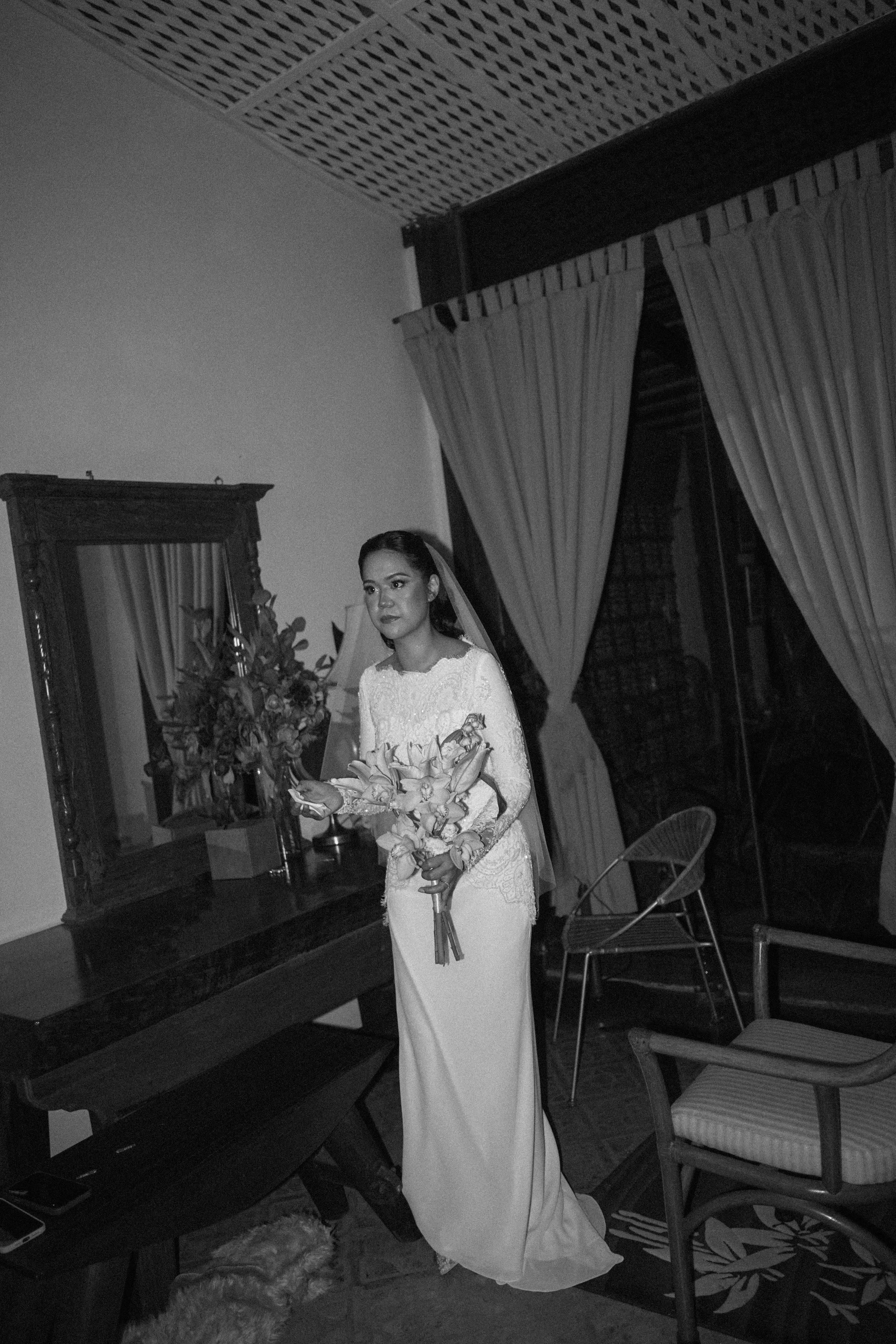 A bride in a white wedding dress holds a bouquet of flowers and stands indoors in front of a mirror and window with curtains, with chairs and furniture visible around.
