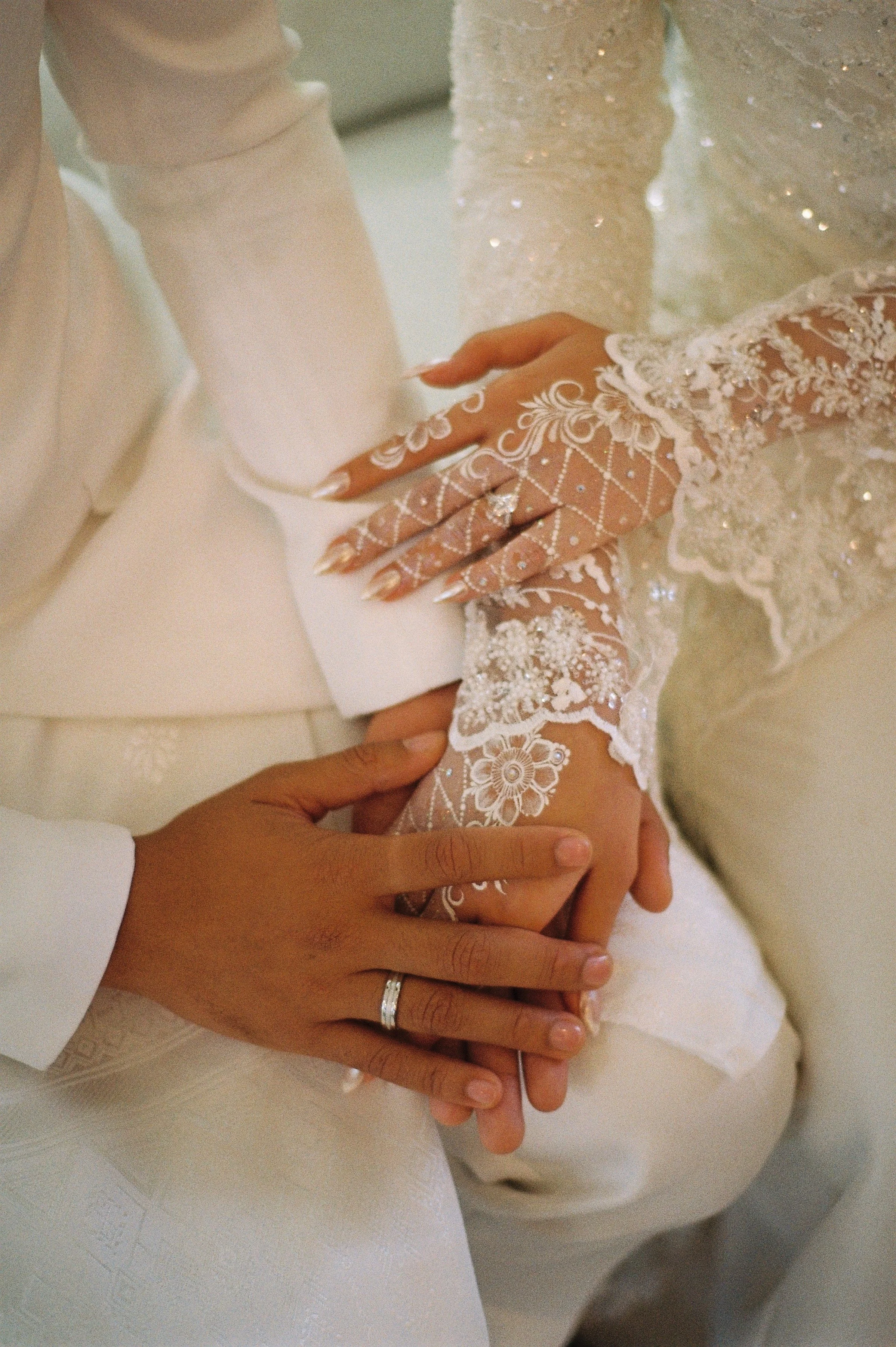 Close-up of a bride and groom's hands intertwined, showing wedding rings; bride's hand is decorated with lace and floral embroidery, groom's hand has a plain band, both on wedding day.