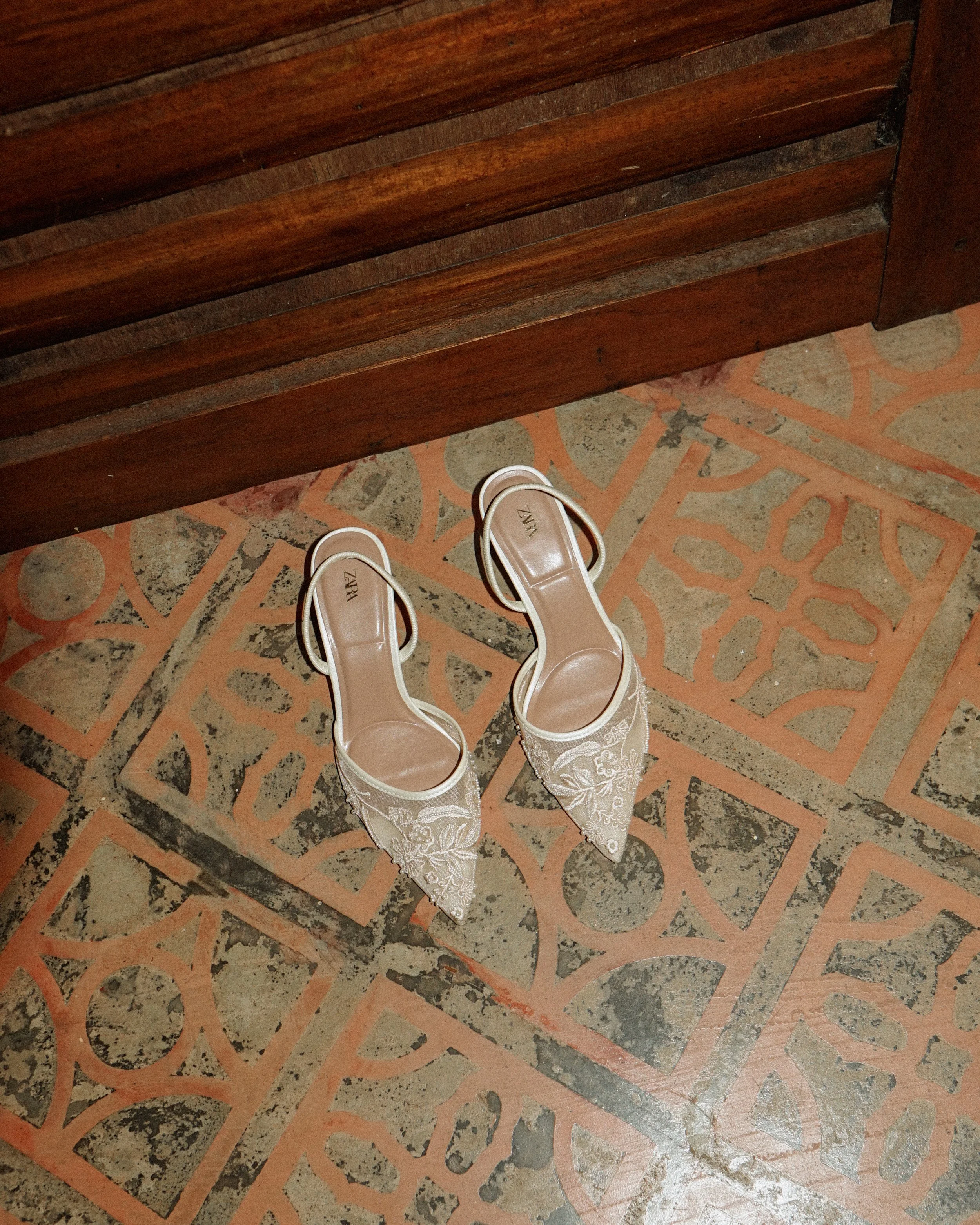 Pair of white embroidered high-heeled shoes on a patterned tile floor next to a wooden bench.
