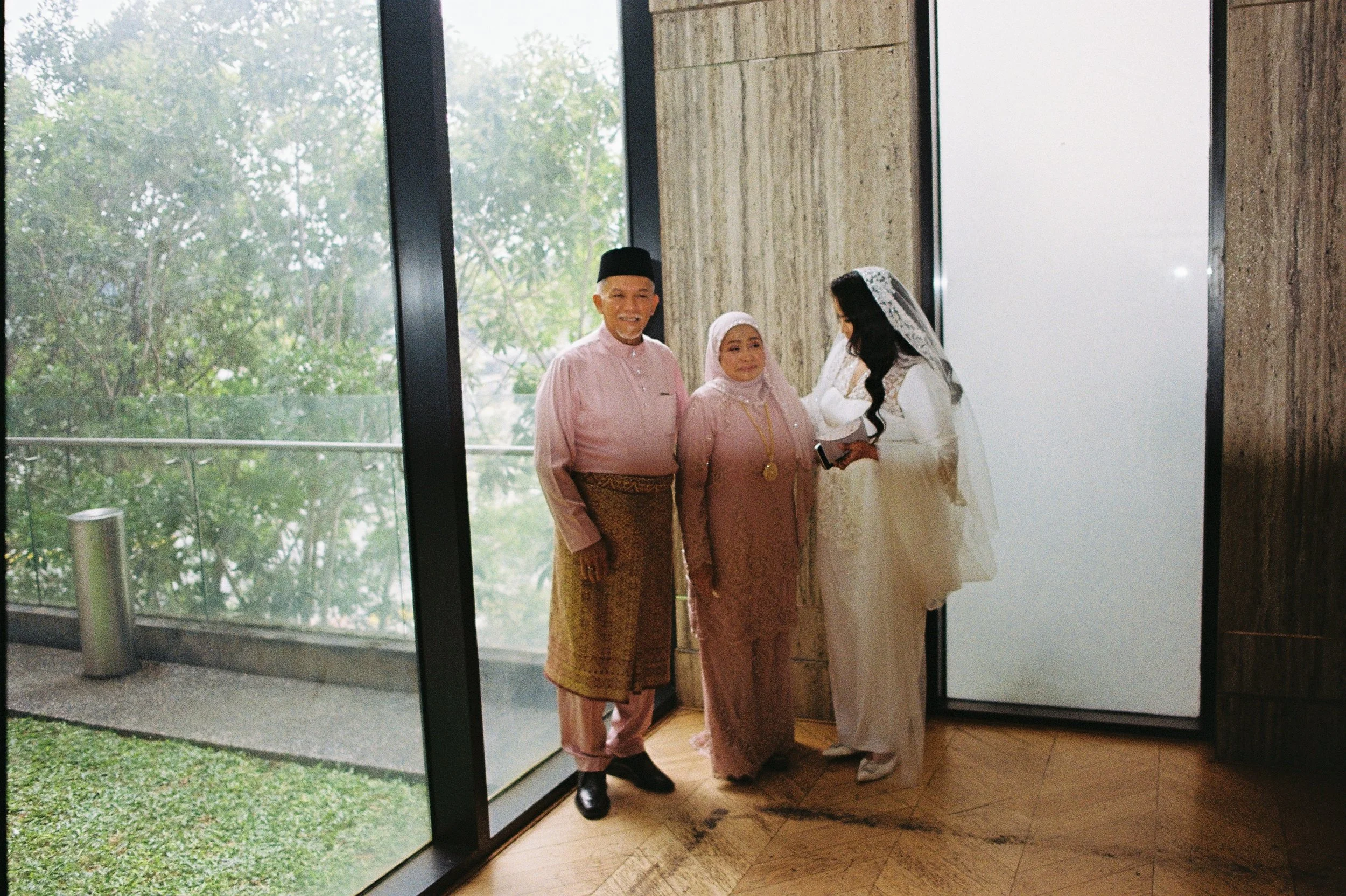 Three people, two women and a man, standing inside near a large window with outdoor greenery visible. The woman on the right is dressed in a wedding gown with a lace veil, and the man and woman on the left are wearing traditional clothing in pink and brown tones. The man is smiling, and the woman beside him is looking at the bride.