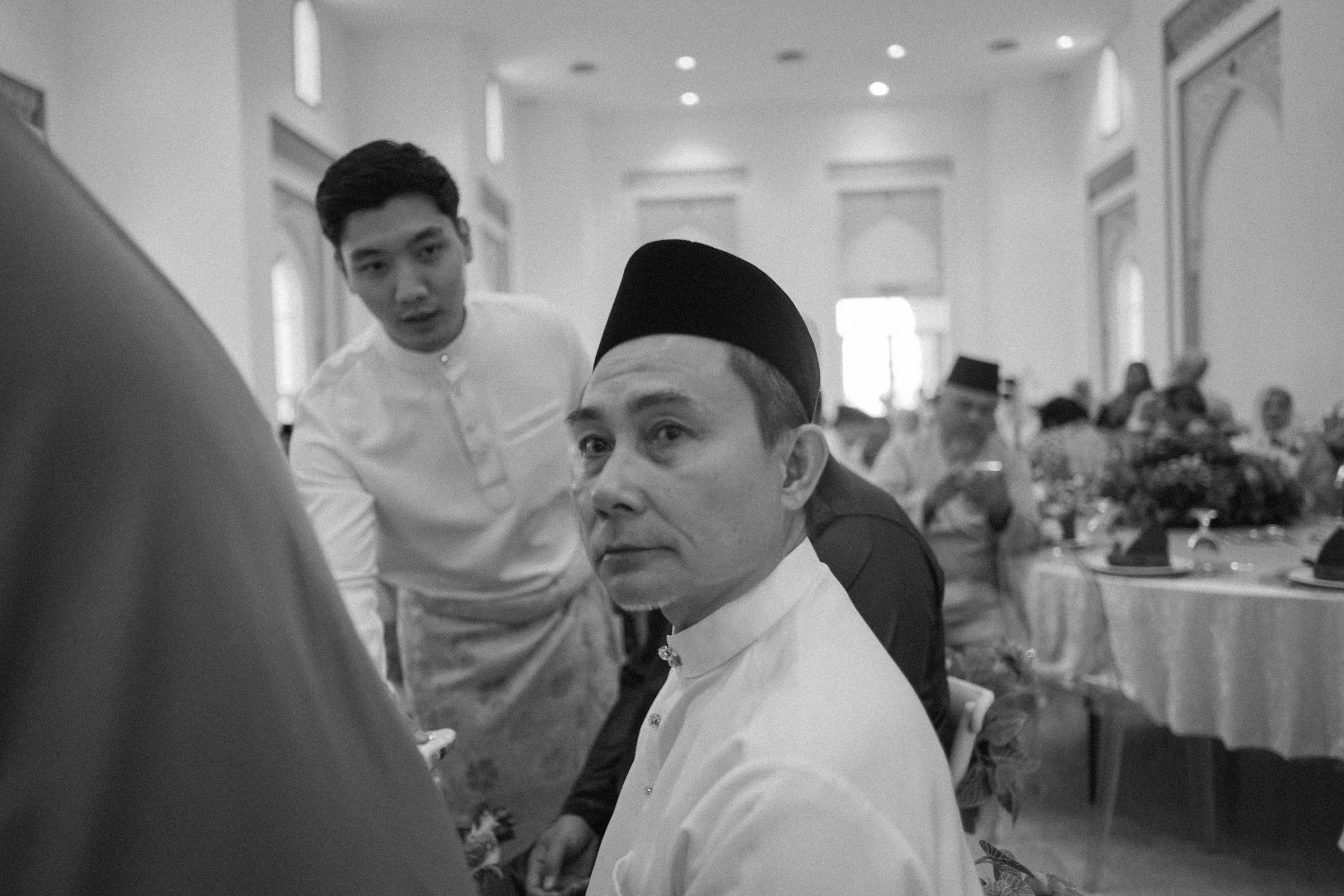 A black-and-white photo of two men at a formal event, one with a traditional cap and the other with a white shirt, in a decorated hall with tables and people in the background.