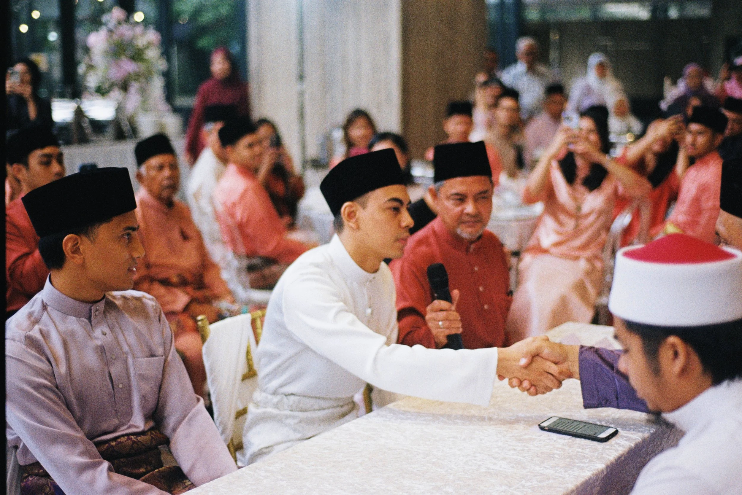 A group of people wearing traditional attire are seated at a table during a cultural event or celebration, with some people in the background.