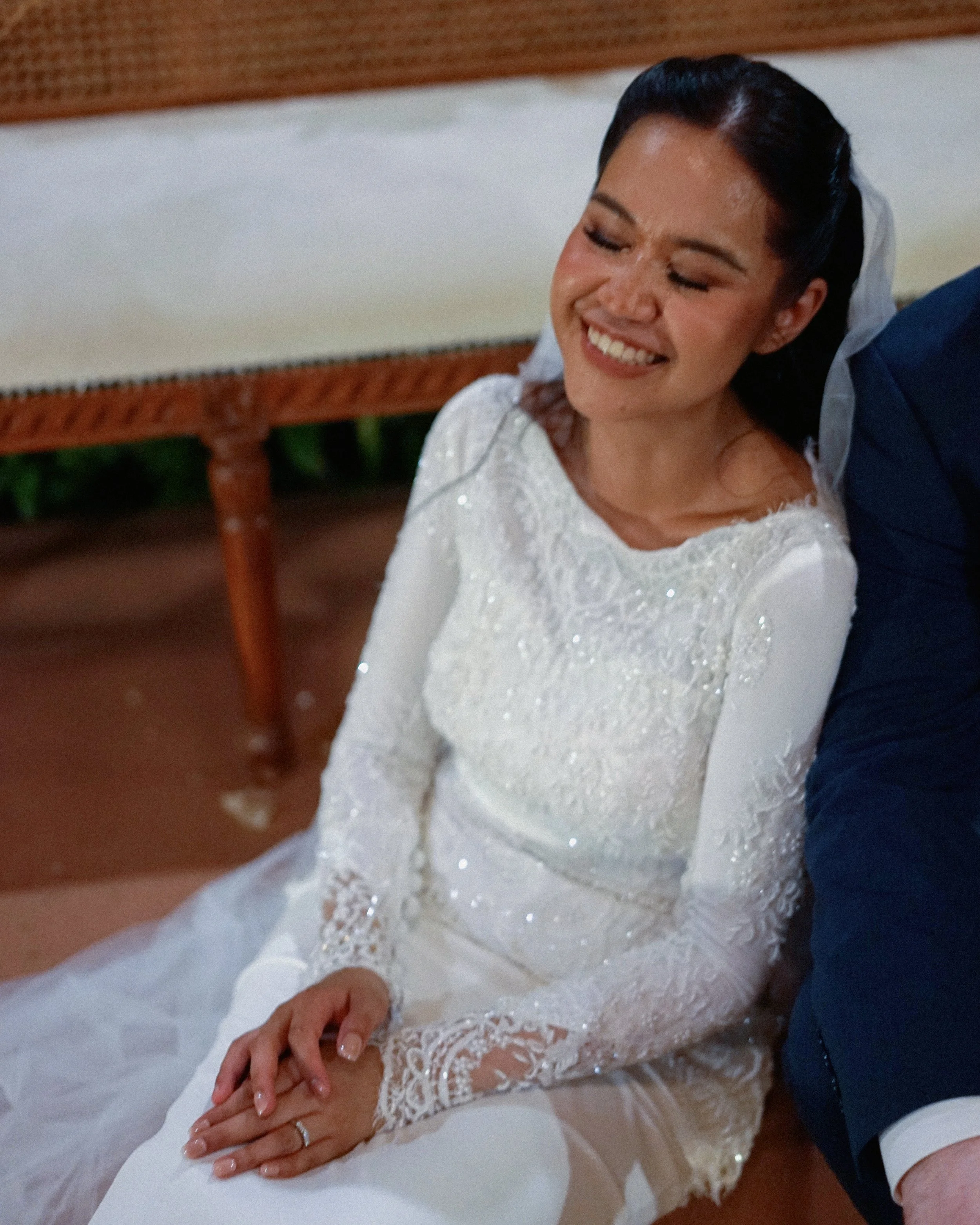 A bride in a white wedding dress resting her hands on her lap while sitting, with a smiling face and closed eyes, accompanied by someone in a blue suit beside her.