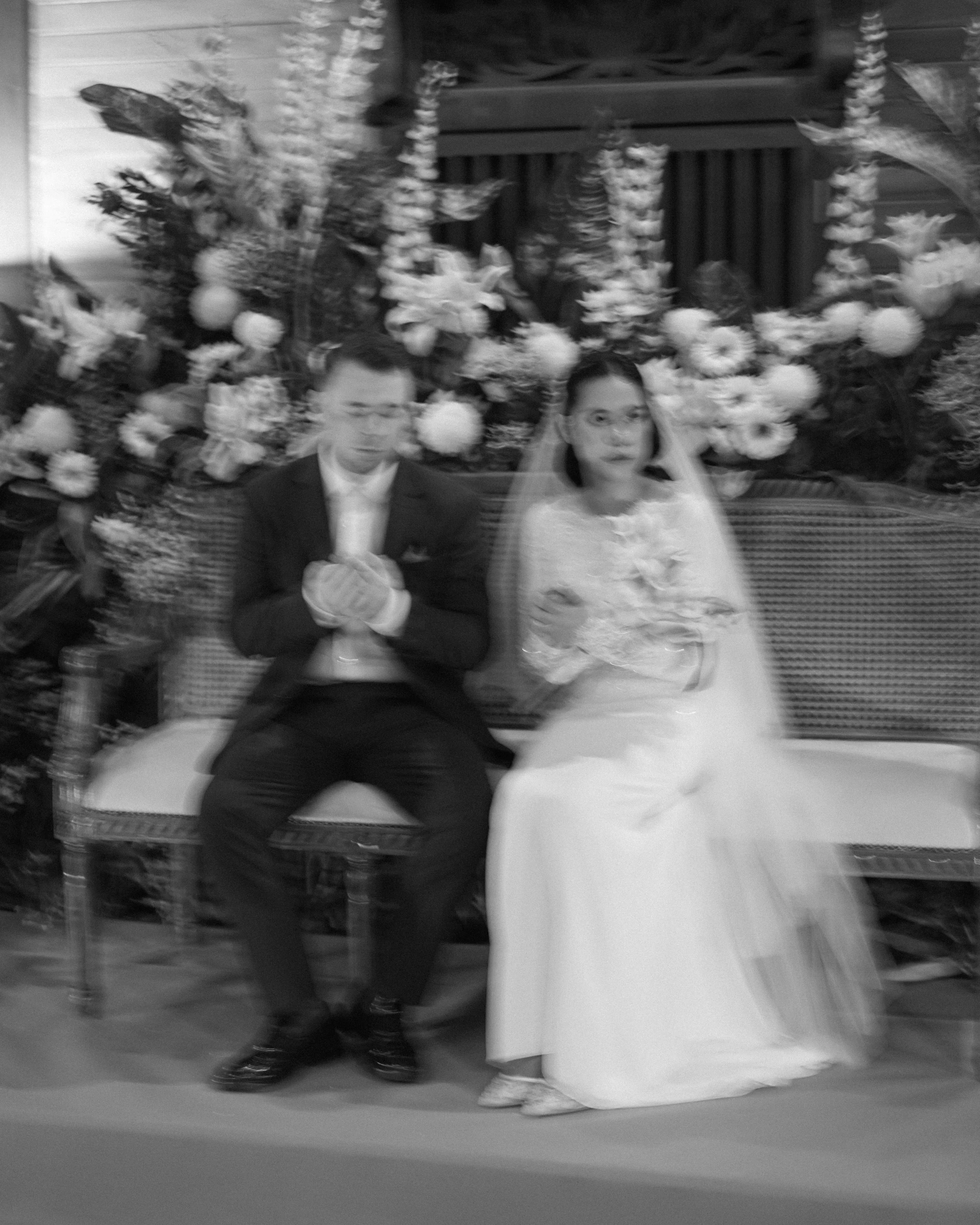 A black and white photo of a bride and groom sitting together on a bench, with a floral wedding backdrop behind them.