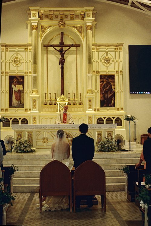 A bride and groom kneeling at the altar during a wedding ceremony in a church, with a priest officiating and religious artwork and a crucifix in the background.