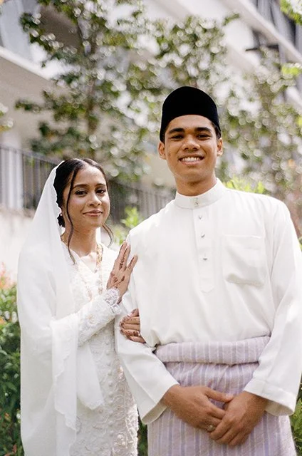 A bride and groom in traditional Malay wedding attire standing outdoors in front of a house. The bride is wearing a white lace dress with a veil, and the groom is dressed in a white shirt, a black songkok, and a striped sarong. They are smiling and holding hands.