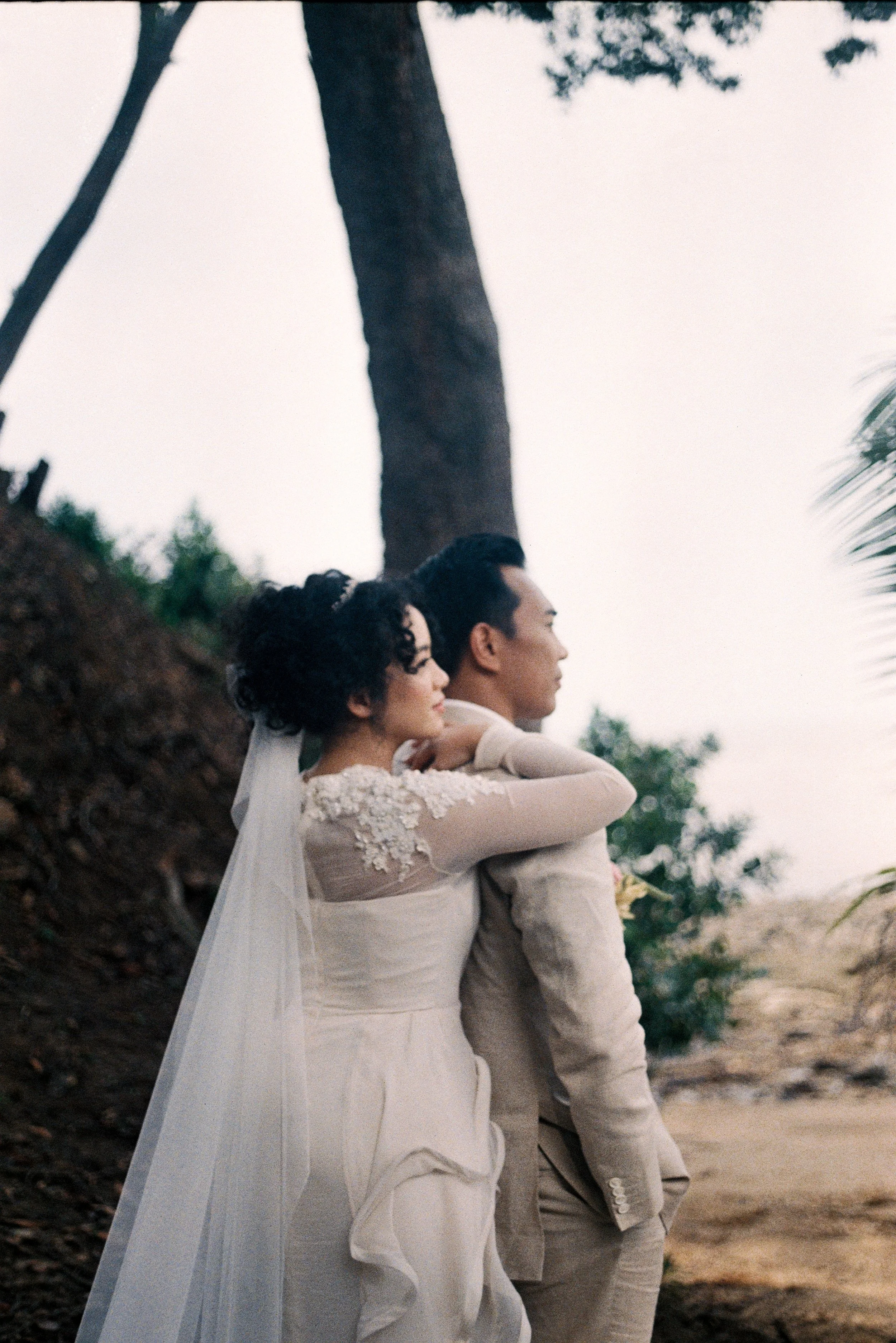 A couple dressed in wedding attire standing outdoors on a beach, facing to the right with their arms crossed, near a tree and sandy terrain.