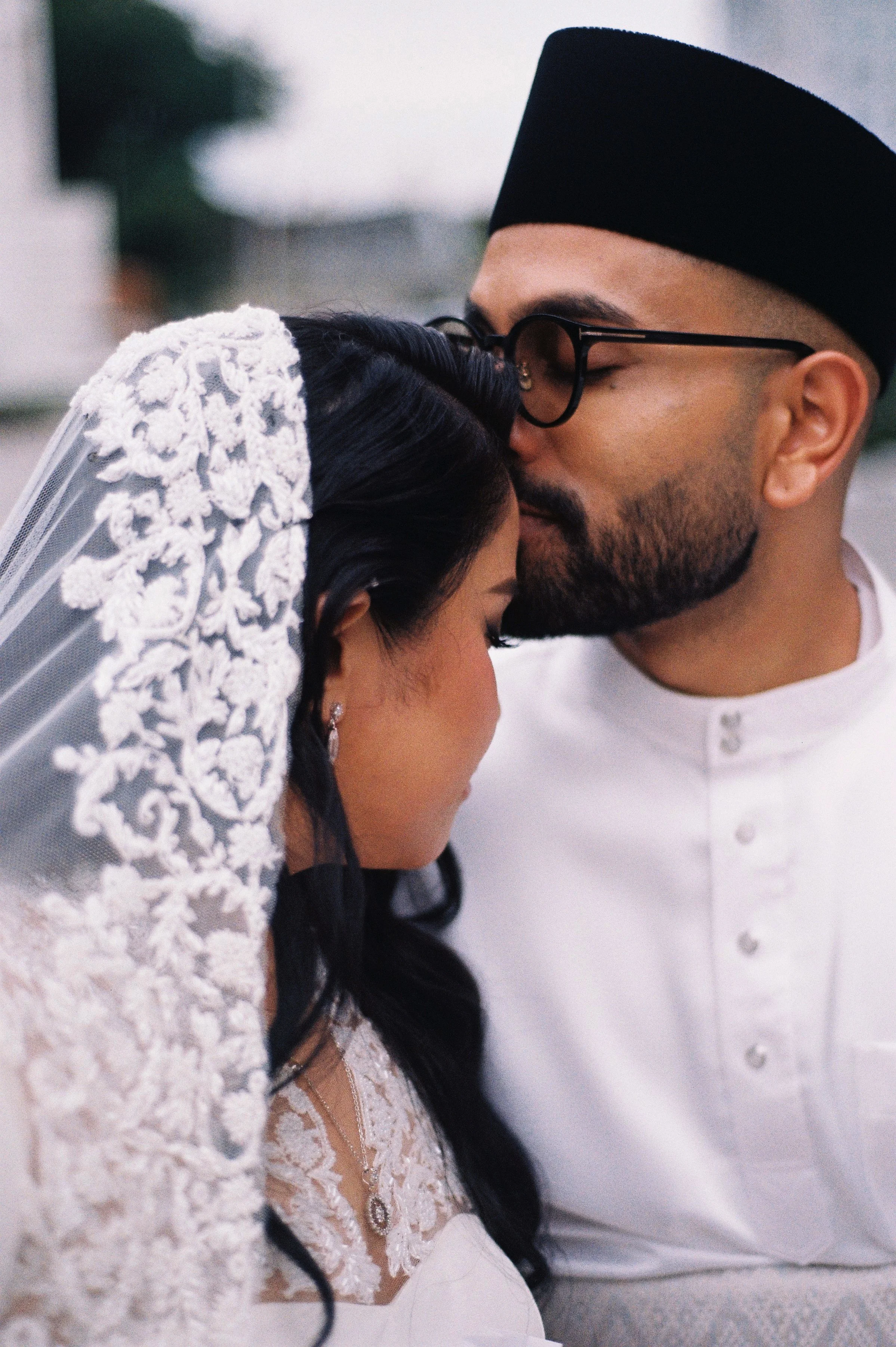 A couple sharing a kiss, with the woman wearing a lace wedding dress and veil, and the man wearing a white shirt and black hat.
