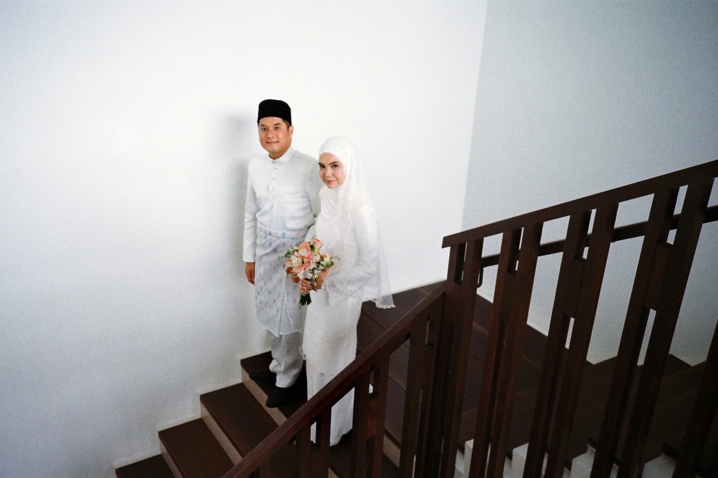 A Muslim couple dressed in traditional white clothing, with the woman holding a bouquet, standing on a staircase against a plain white wall.