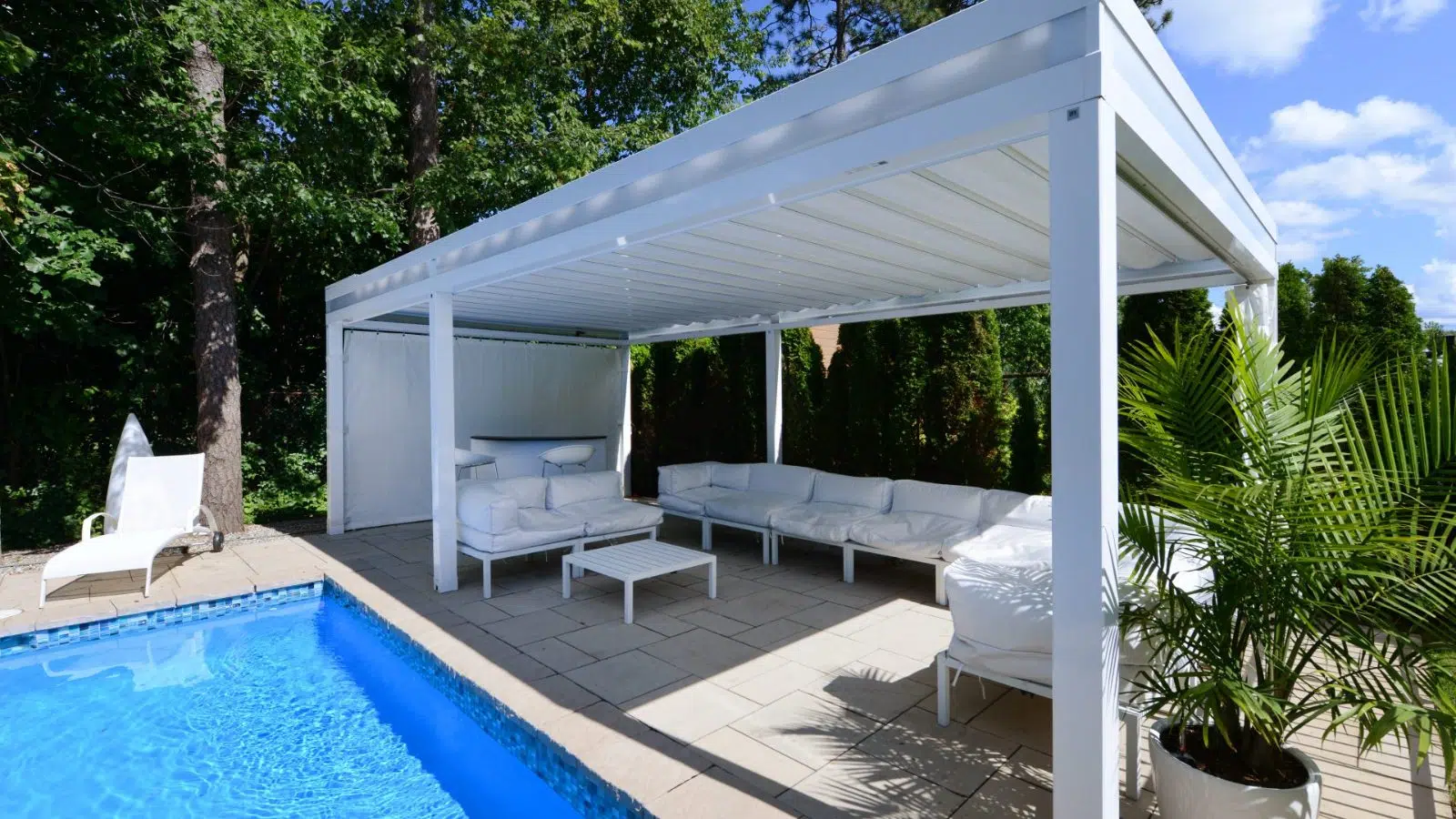 Outdoor patio with a swimming pool, white seating under a white pergola, potted plants, and lush greenery in the background.