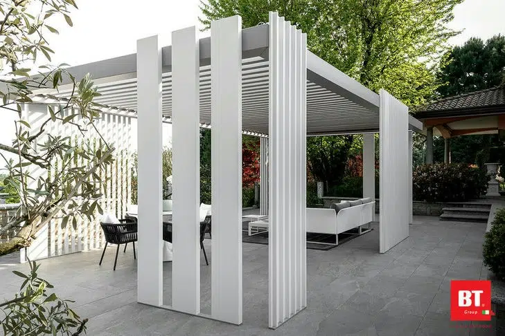 Modern outdoor patio with a white pergola and furniture including a white sofa and dining table with black chairs, surrounded by greenery.