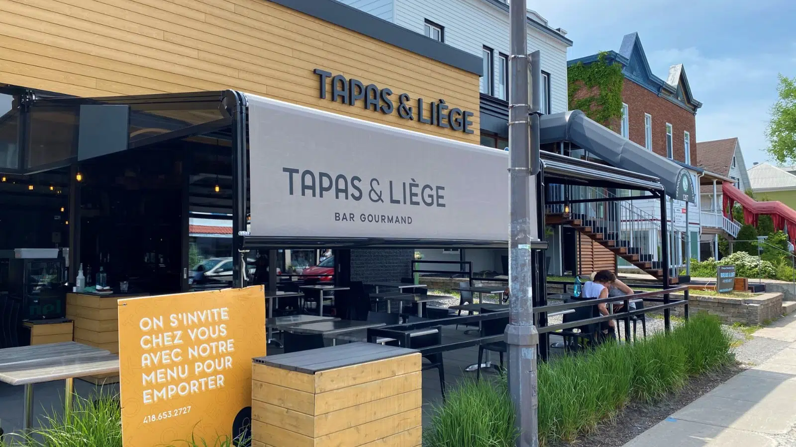 Exterior view of a tapas and liquor restaurant with a sign that reads 'Tapas & Liège' and an outdoor seating area. There's a yellow standee with French text inviting customers to dine and take out, and a couple of people seated outside. Buildings and