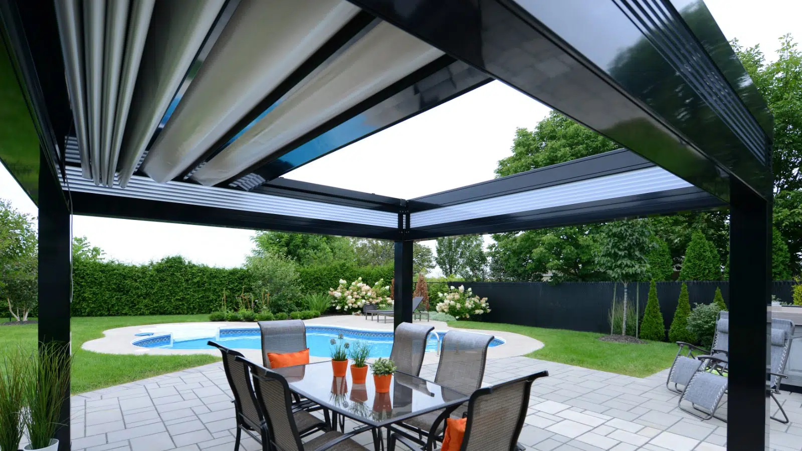 View of a backyard patio with a black pergola, outdoor dining table with chairs, swimming pool, and lush green landscaping.