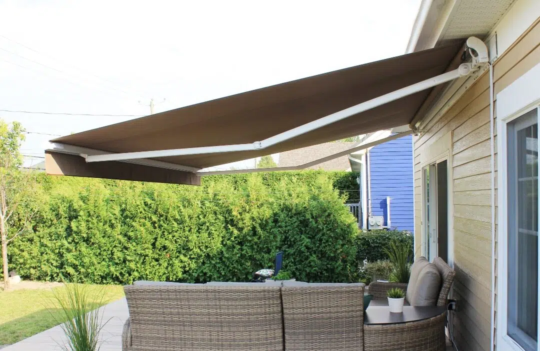 Covered outdoor patio area with a wicker outdoor sofa, potted plants, and an extendable brown fabric awning attached to a yellow house.