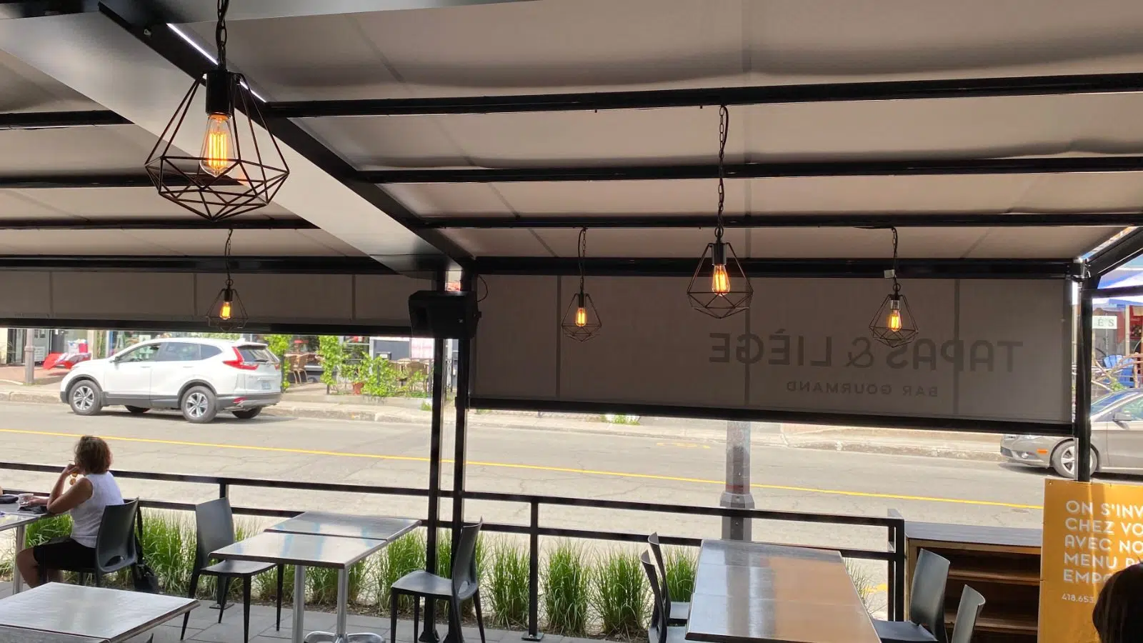 Interior of a cafe with white tables and black chairs, a woman sitting at a table near the window, and hanging geometric pendant lights. Outside view of parked cars and storefronts.