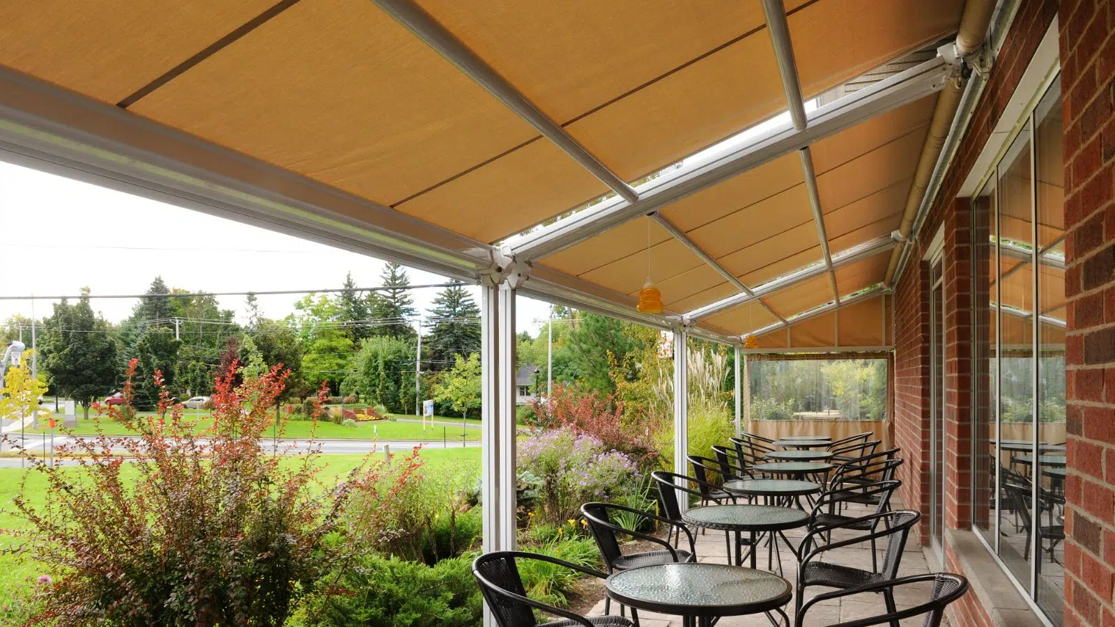 Outdoor patio with black metal tables and chairs under a brown awning, adjacent to a brick building, overlooking a green lawn with trees and shrubbery.