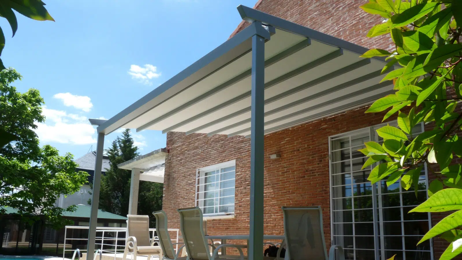 Backyard patio with a metal awning, brick house exterior, chairs, trees, and blue sky.