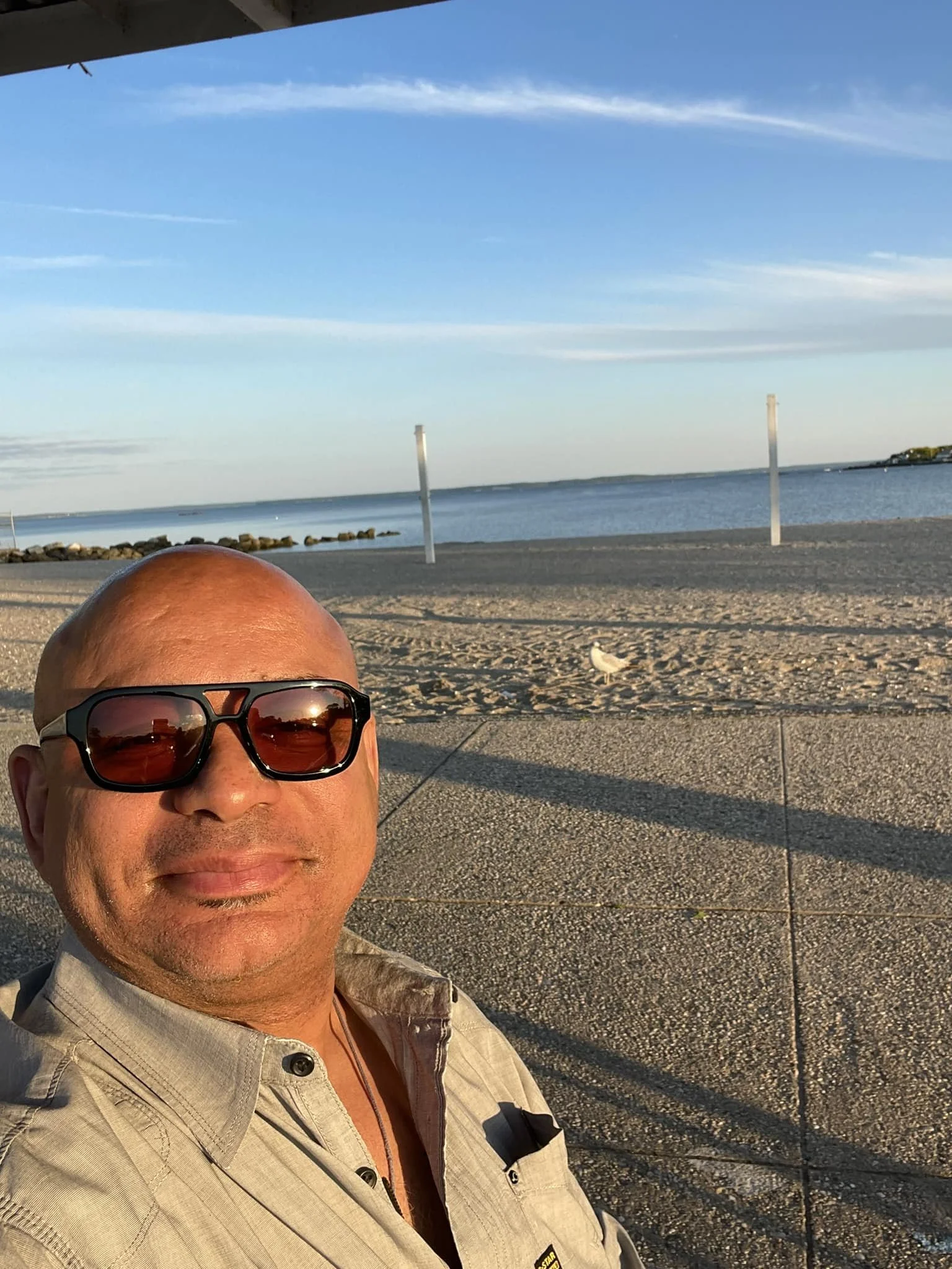 A man wearing sunglasses takes a selfie at a beach during sunset, with sand, water, and a blue sky with clouds in the background.