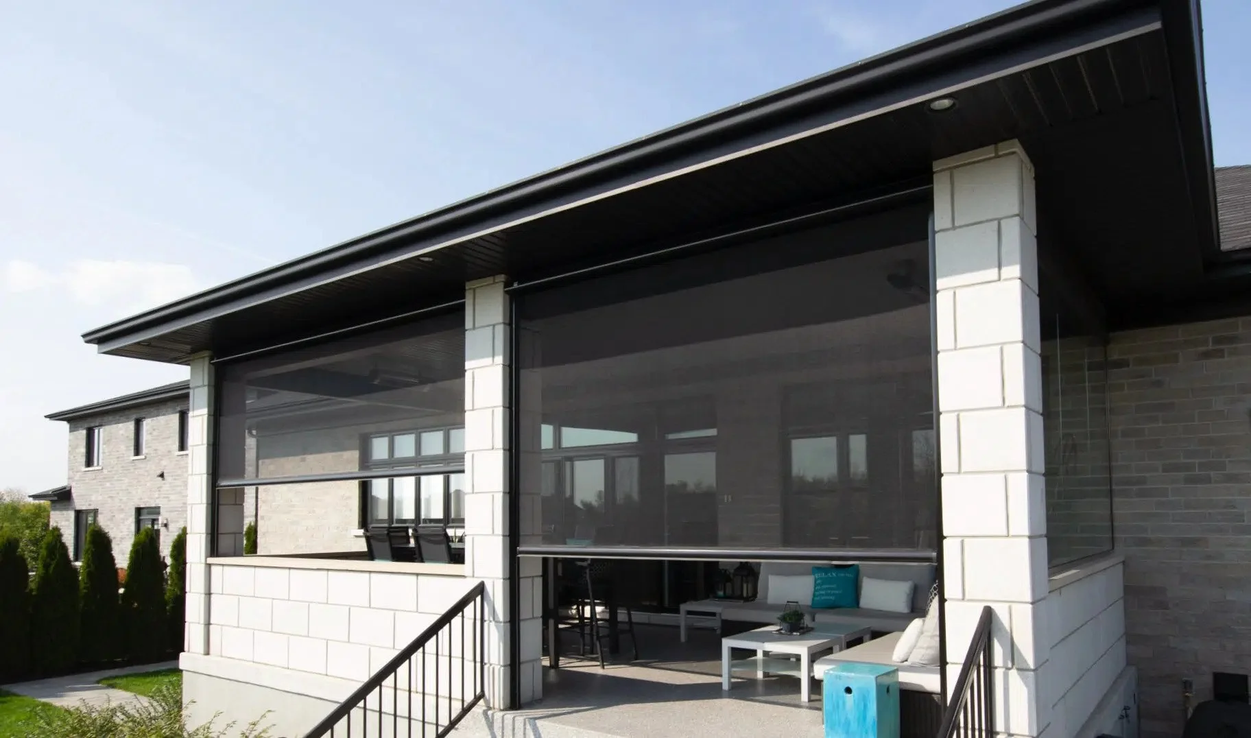 Back porch of a modern house with outdoor furniture, including a white table and a white sofa with a blue pillow, shaded by a black screen canopy, with brick and siding exterior walls, stairs with black railing, and a yard with green bushes.