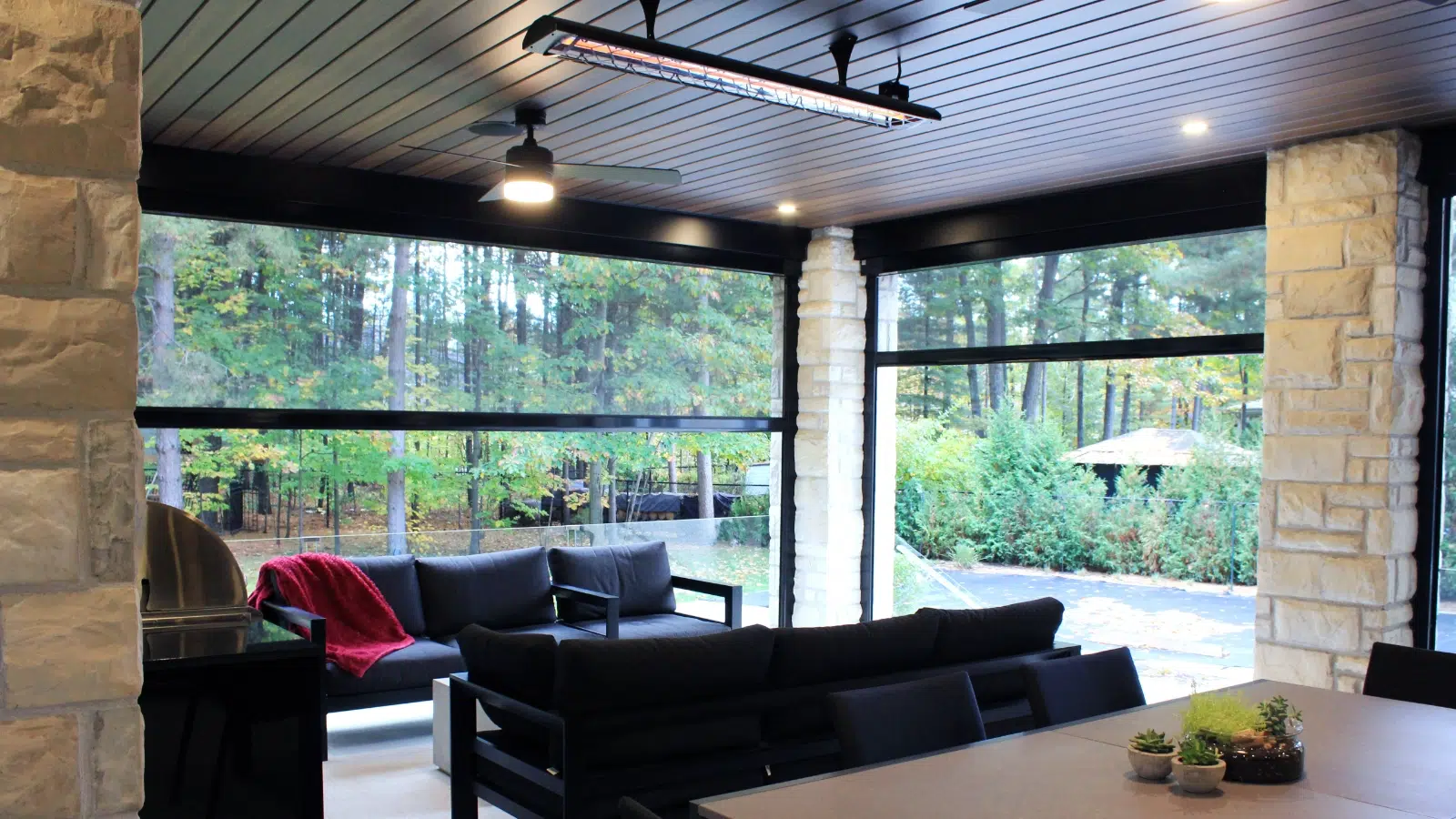 Indoor living area with large windows showing a wooded outdoor scene, black sofas with a red blanket, a patio outside, and a dining table with small potted plants.