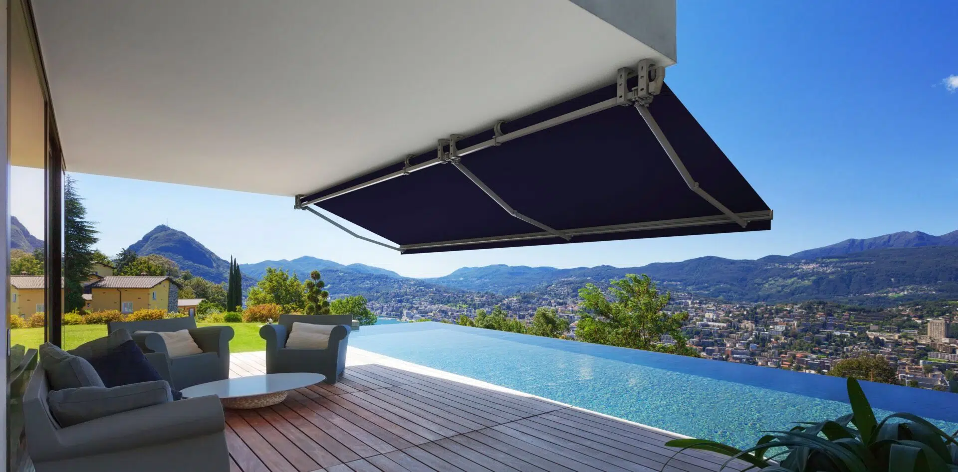 Modern outdoor patio with a view of mountains and cityscape, featuring a covered space with comfortable seating, a wooden deck, and an infinity pool.