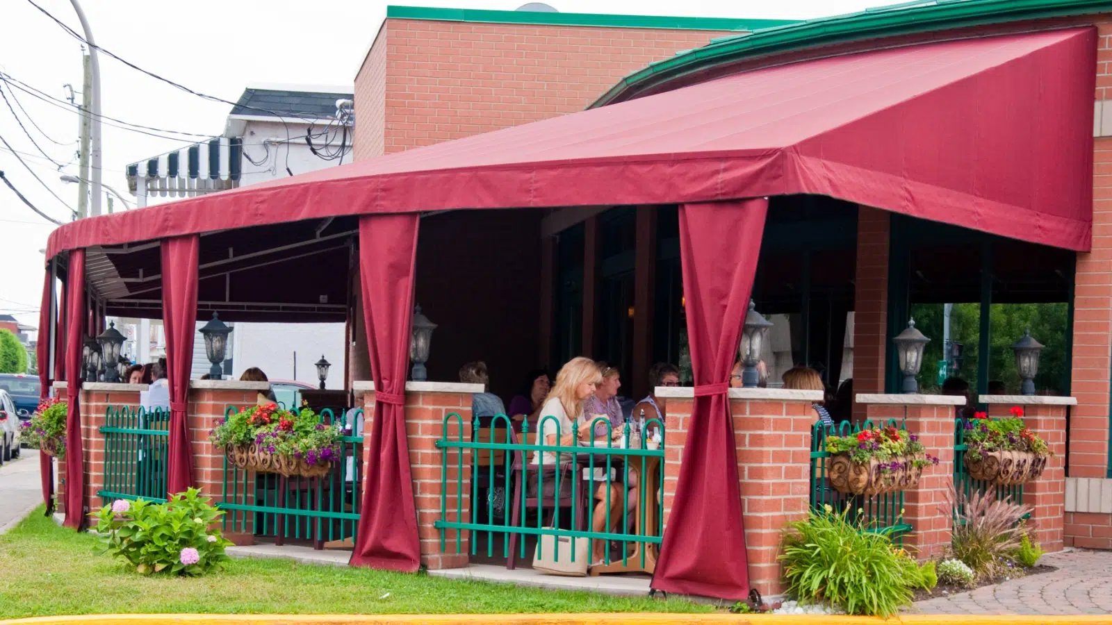 People sitting and dining under a red canopy at an outdoor restaurant with brick walls, potted flowers, and hanging lanterns.