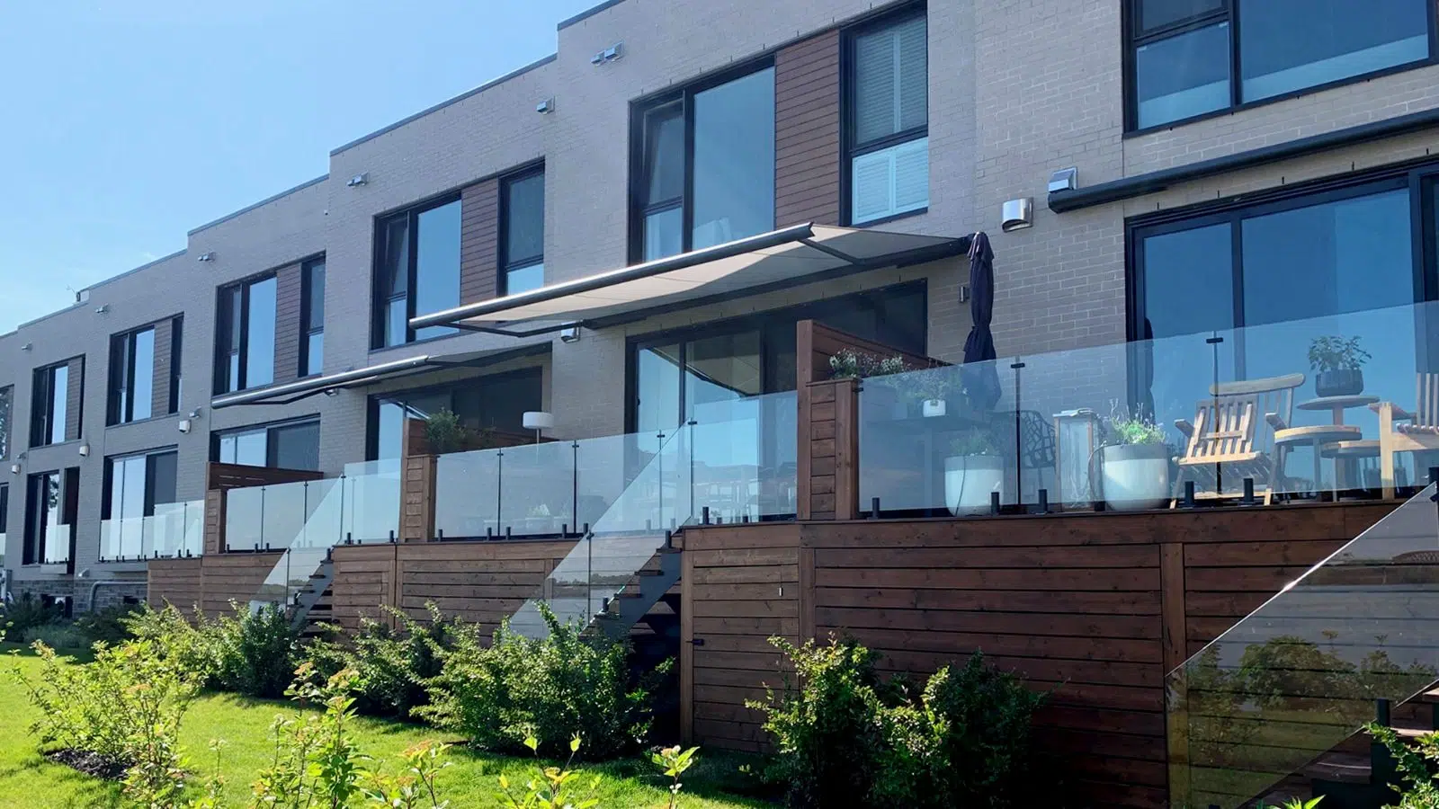 Modern apartment building with glass balconies, wooden fencing, outdoor furniture, and greenery in front under a clear sky.