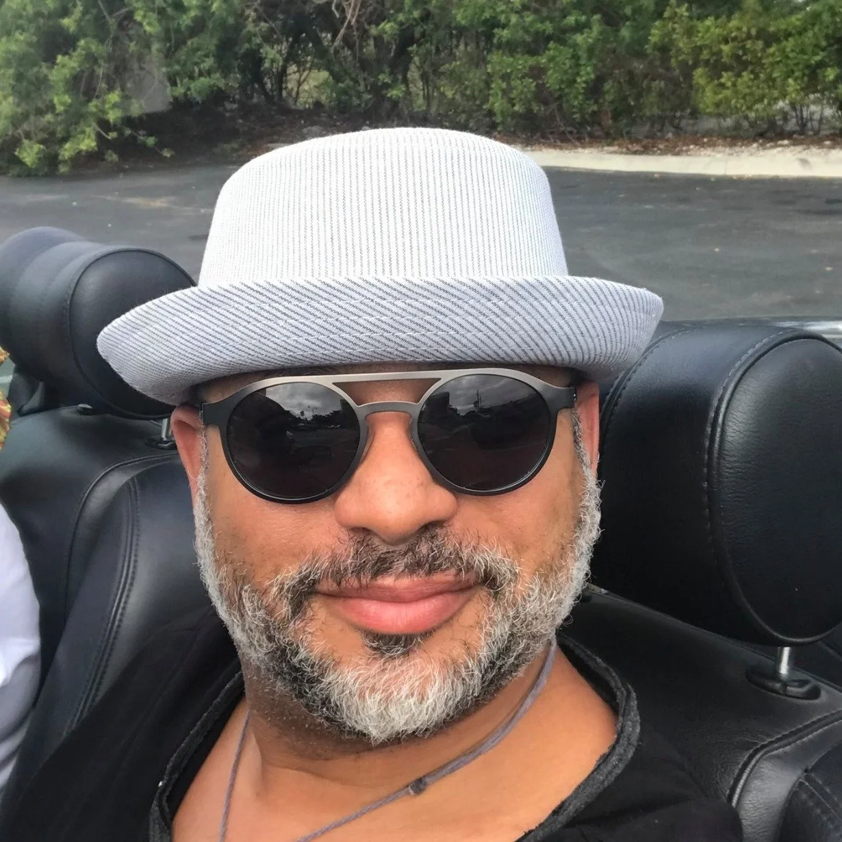 A man with sunglasses, a gray beard, and a light-colored hat takes a selfie inside a convertible car. There are green bushes and a pavement visible in the background.