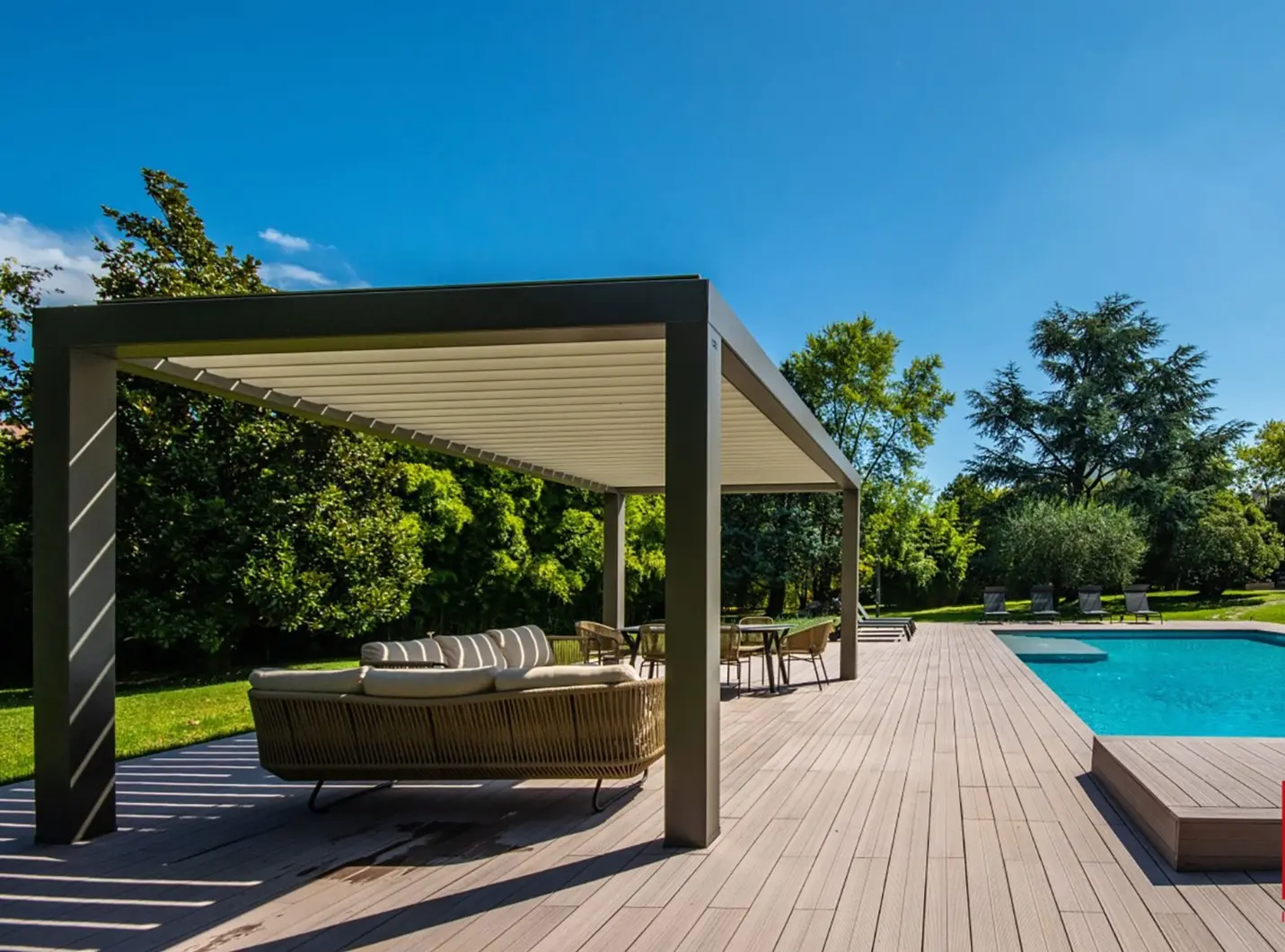 Outdoor poolside area with a pergola, lounge seating, and swimming pool surrounded by green trees under a clear blue sky.