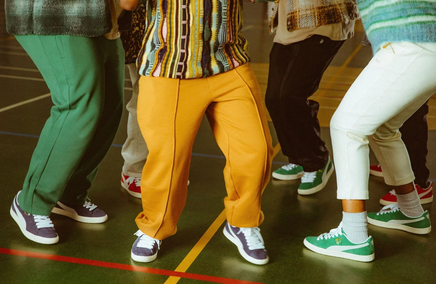 Close-up of people dancing, showing their colorful pants and sneakers. The setting appears to be a gym or indoor sports court.