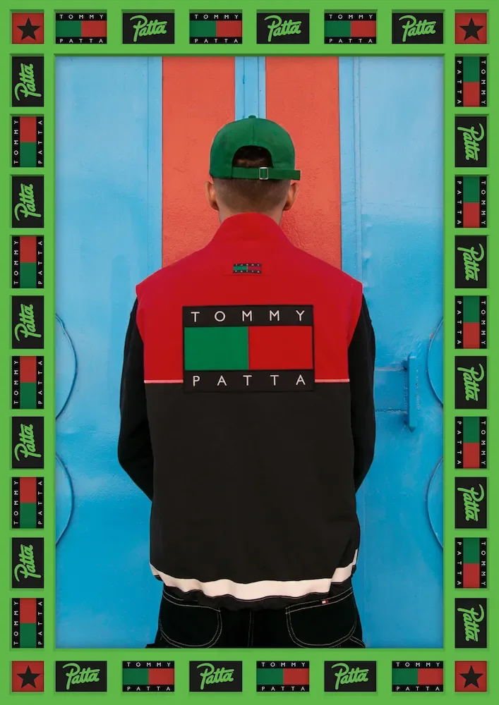 A person stands with their back to the camera in front of a vibrant blue and orange wall, wearing a green cap and a black and red jacket with a Tommy Hilfiger logo on the back.