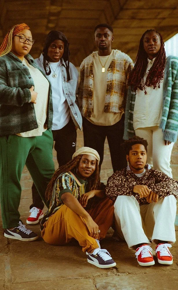 Group of six young adults posing indoors with a wooden ceiling background, some sitting on the ground and others standing, dressed casually in colorful, patterned clothing.