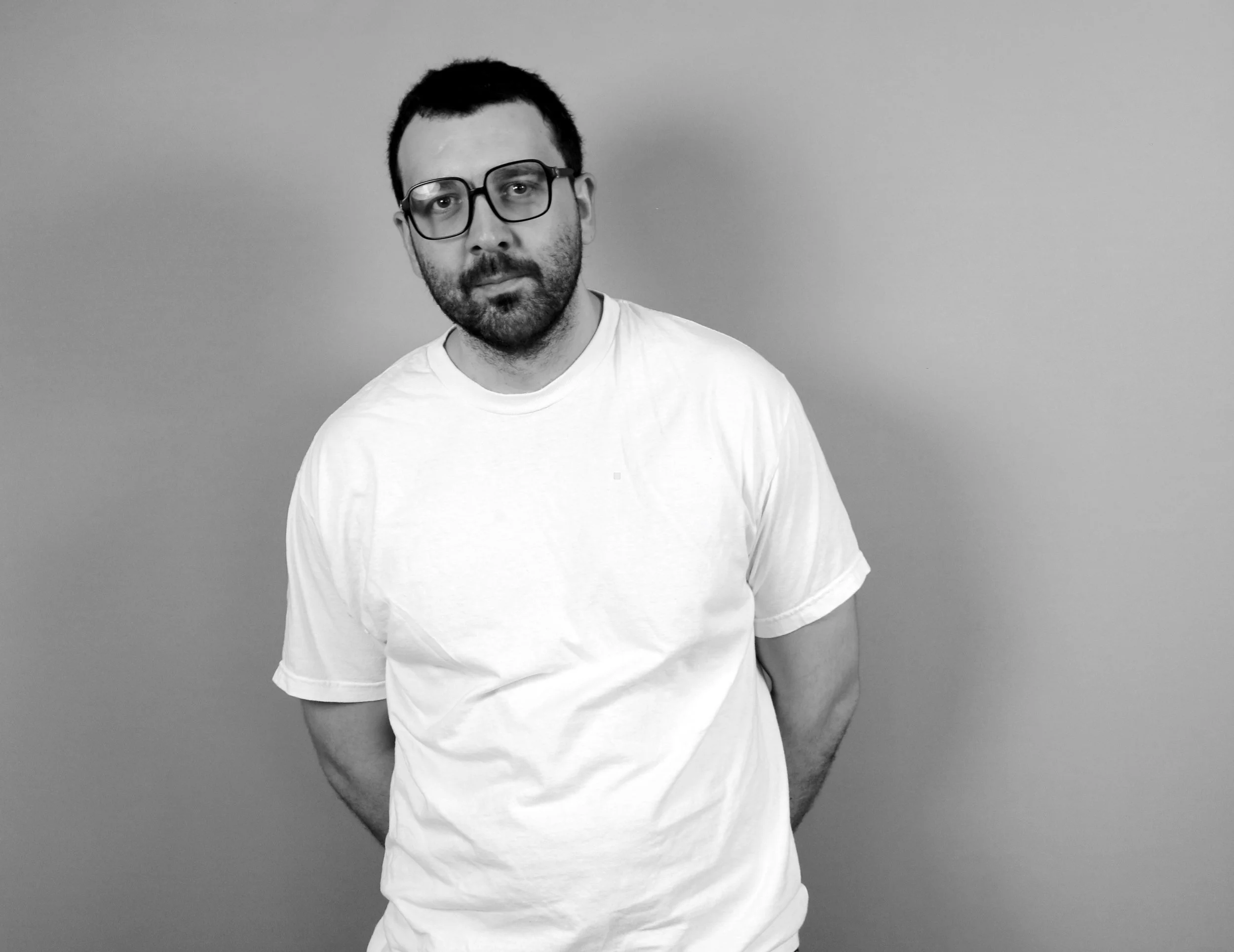 A man with glasses, a beard, and short dark hair wearing a white t-shirt posing against a plain background.